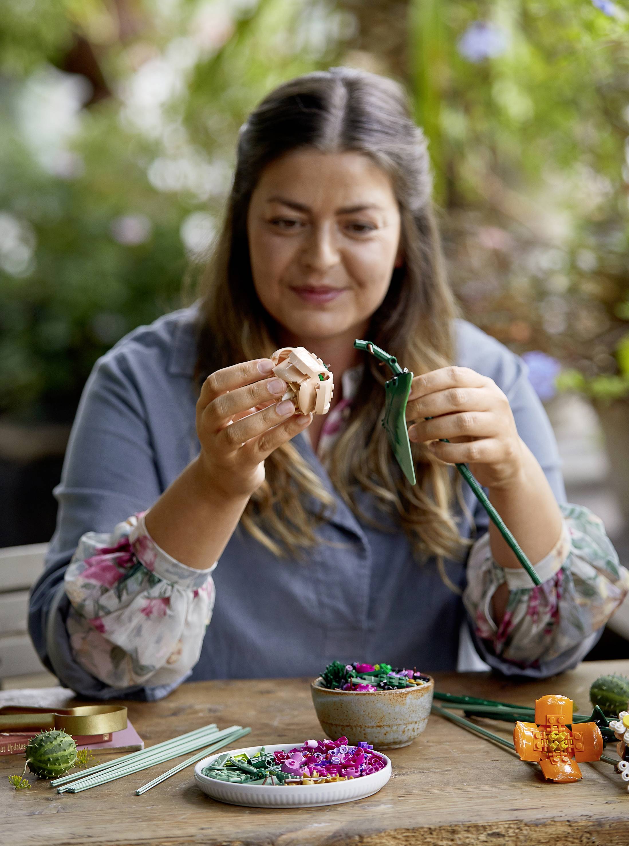 Vrouw maakt bloemstuk van kunstmaterialen aan een tafel met toebehoren. Achtergrond toont onscherpe planten.