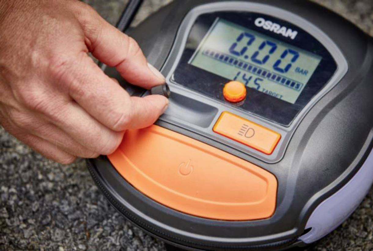 Een hand bedient een digitaal bandenspanningsmeetapparaat van Osram, dat 0,0 bar aangeeft op een asfaltoppervlak.