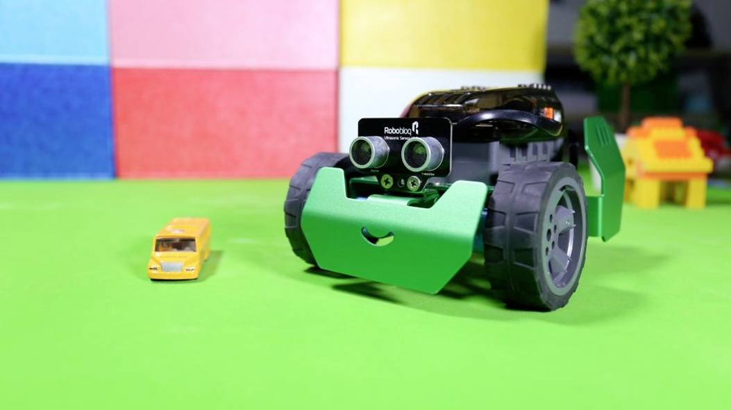Speelgoedrobot met een vrolijk gezicht rijdt op een groene ondergrond naast een kleine gele speelgoedauto. Kleurrijke muur op de achtergrond.