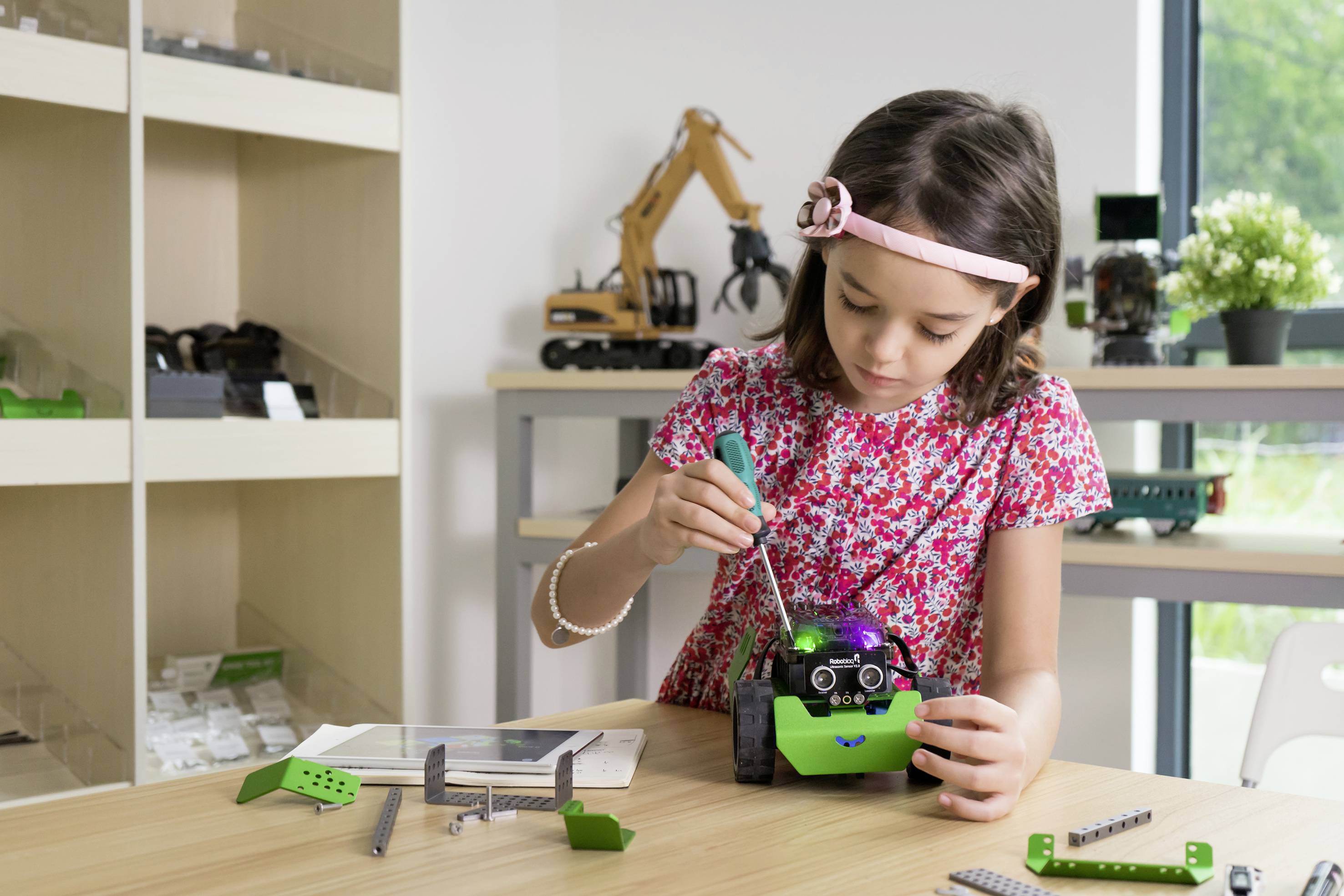 Een meisje repareert een kleine robot met een schroevendraaier aan een tafel in een ruimte vol speelgoed en modellen.