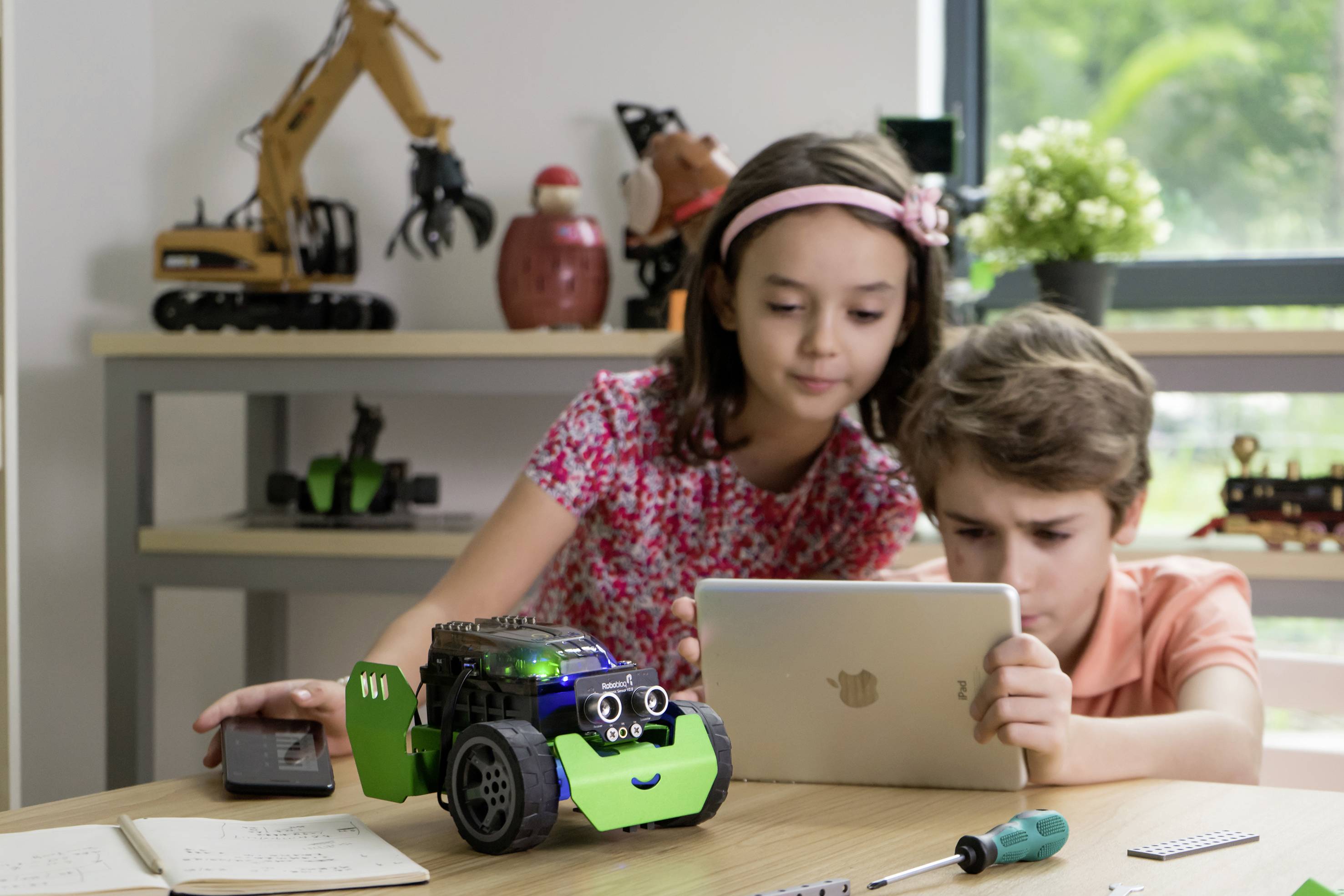 Een meisje en een jongen interageren met een tablet terwijl ze een kleine, robotische speelgoedauto programmeren. Op de achtergrond zijn leermaterialen te zien die wijzen op techniek.