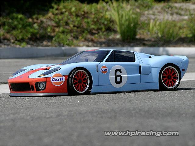 Blauw en oranje modelauto in Gulf-design met nummer 6, geparkeerd op asfalt, omgeven door groene vegetatie op de achtergrond.