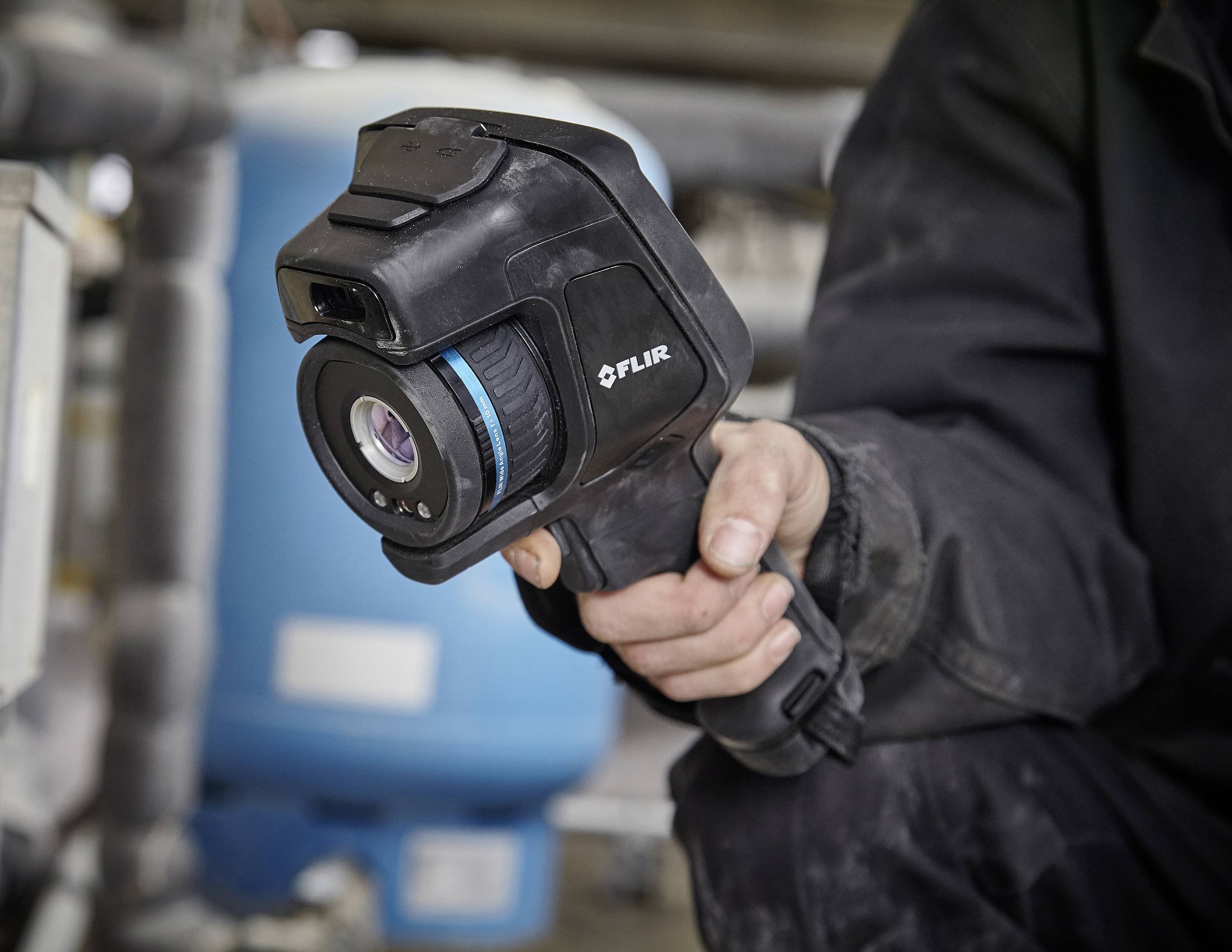 Een persoon houdt een FLIR-warmtebeeldcamera in de hand, omgeven door technische apparaten.