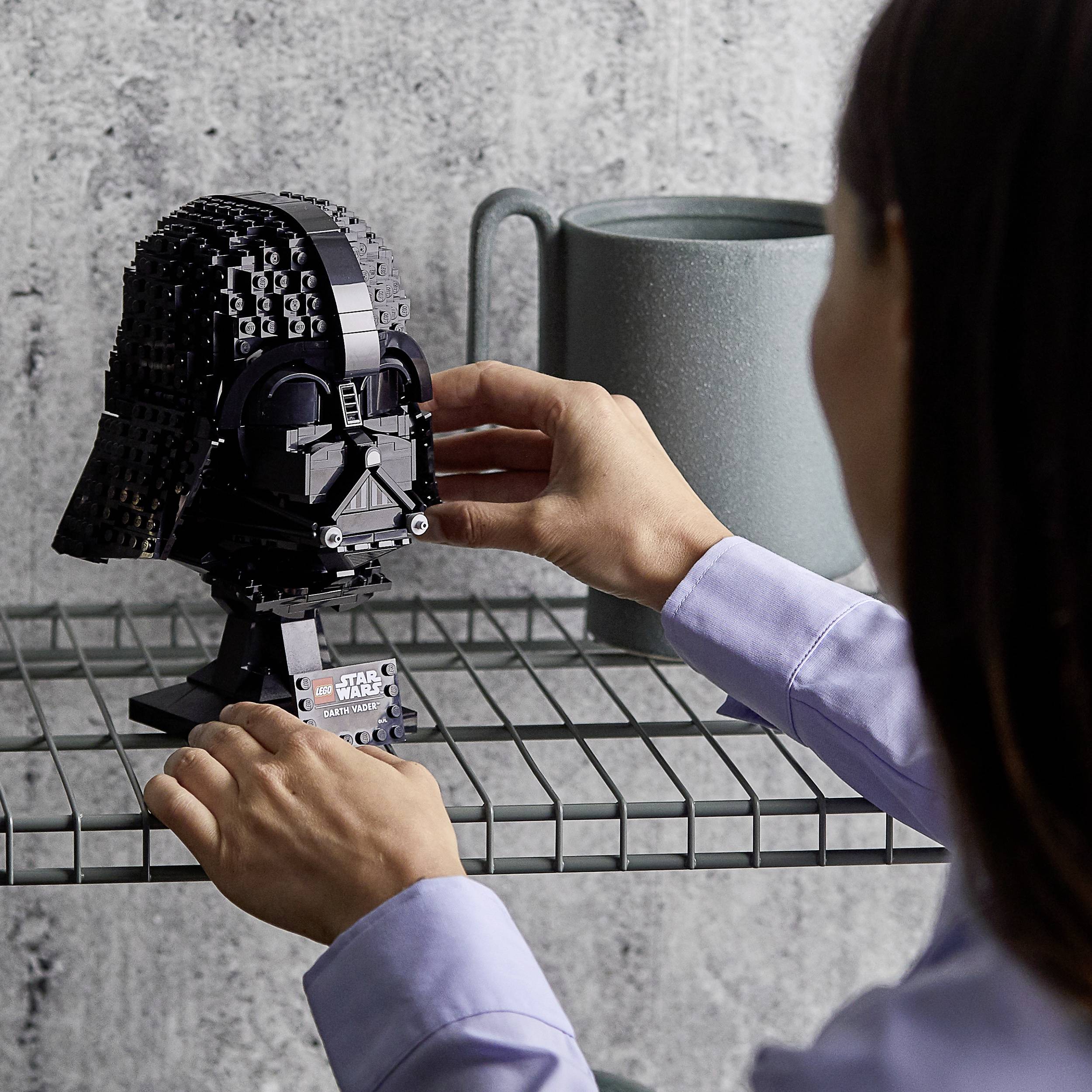 Een persoon plaatst een 'Star Wars' Lego-model van een zwarte helm op een metalen rek. Op de achtergrond staat een grijze mok.