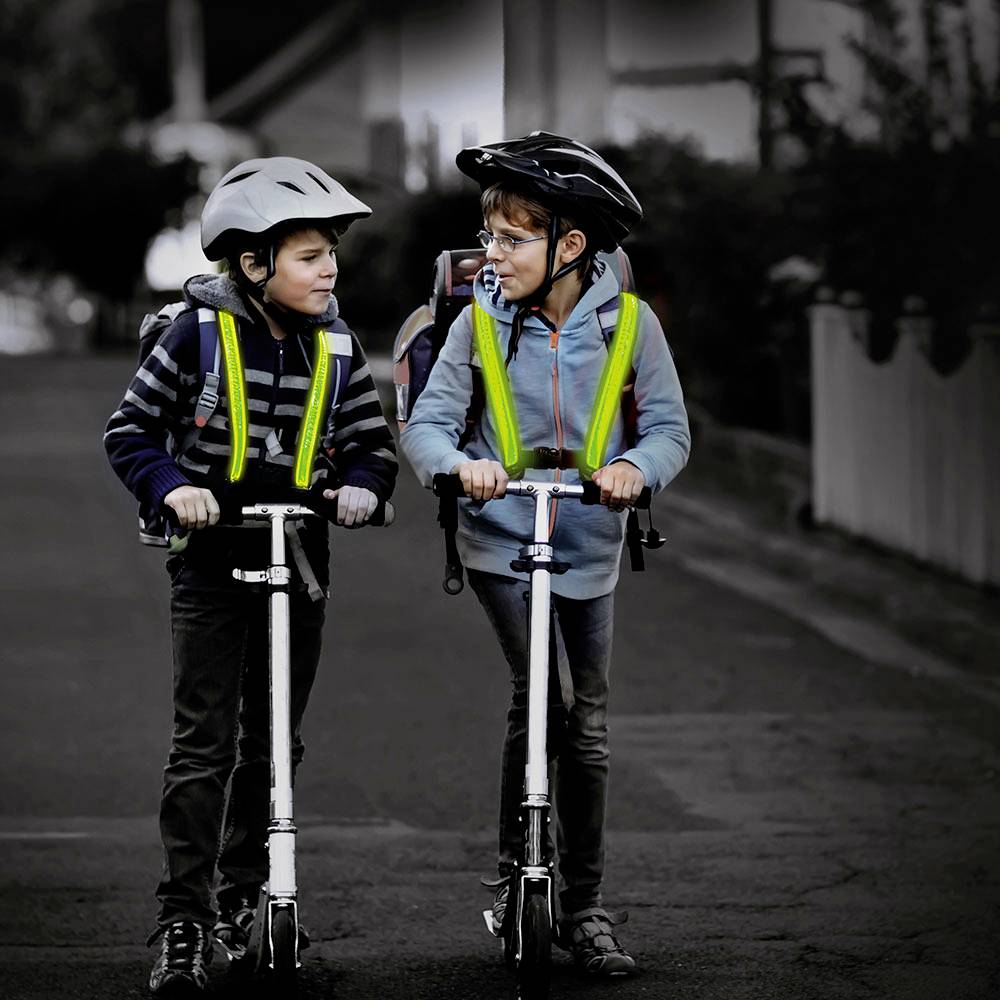 Twee kinderen met helmen en reflecterende hesjes rijden op steps langs een straat. Ze praten met elkaar en dragen rugzakken.