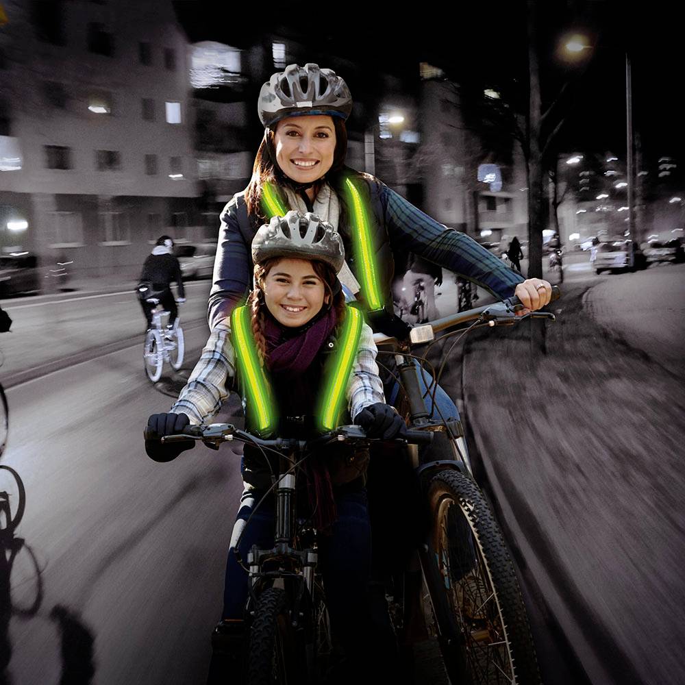Een vrouw en een kind, beiden met helmen, rijden 's avonds samen op een fiets. Hun reflecterende hesjes lichten op in het donker.
