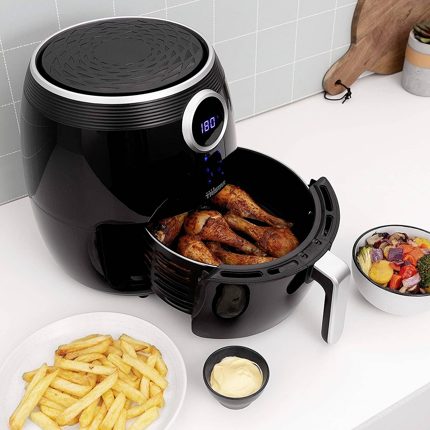 Airfryer in keuken met geopende lade vol gebakken kippenpoten. Ernaast friet met dip en groentesalade.