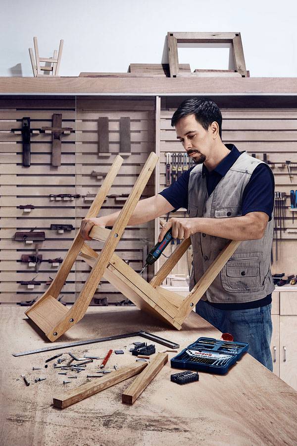 Een man monteert een houten stoel in een werkplaats; omringd door gereedschappen zoals schroevendraaiers en schroeven, met houten rekken op de achtergrond.