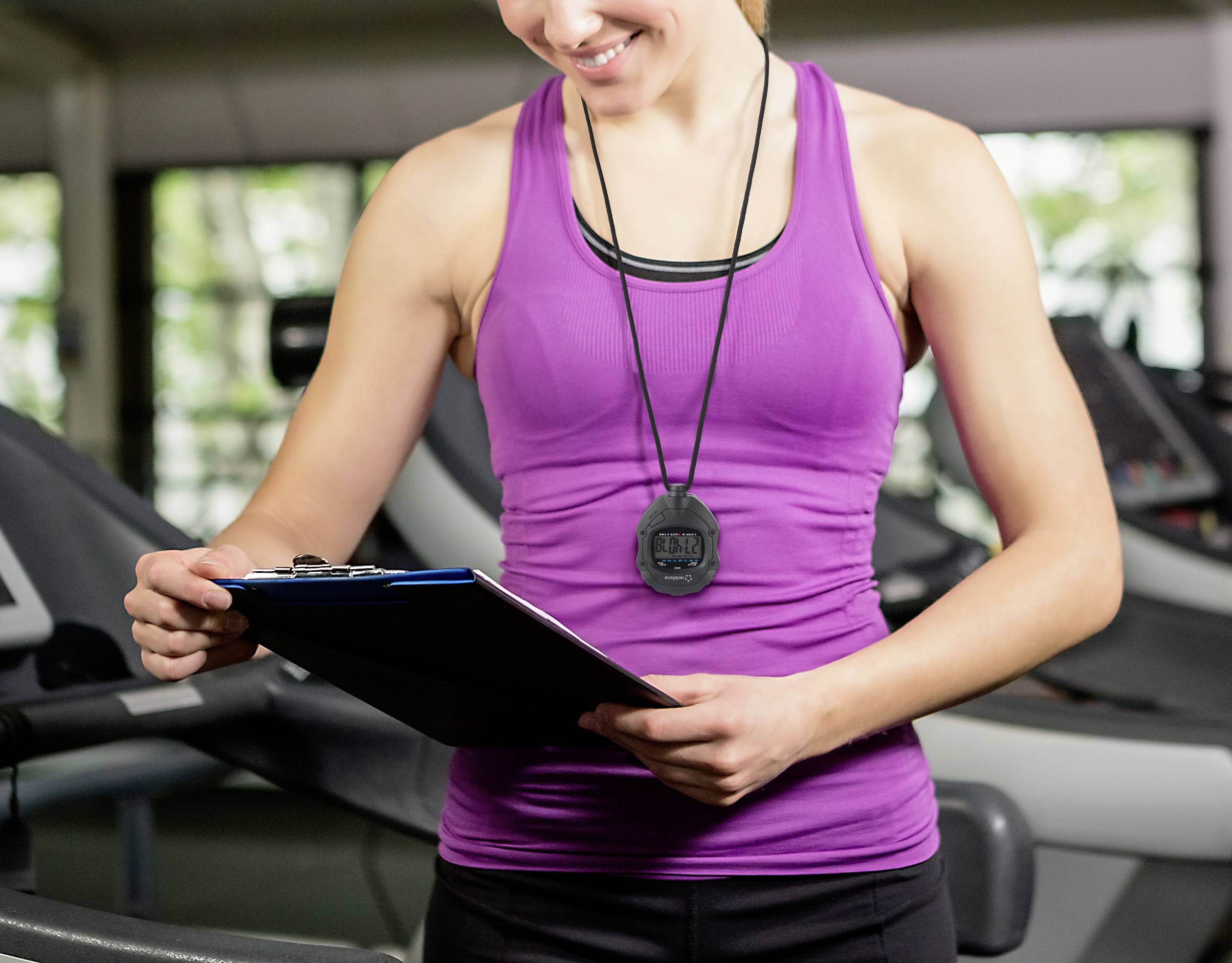 Een vrouw in de sportschool draagt een paars sporttopje en houdt een klembord vast. Op de achtergrond zijn fitnessapparaten zichtbaar.