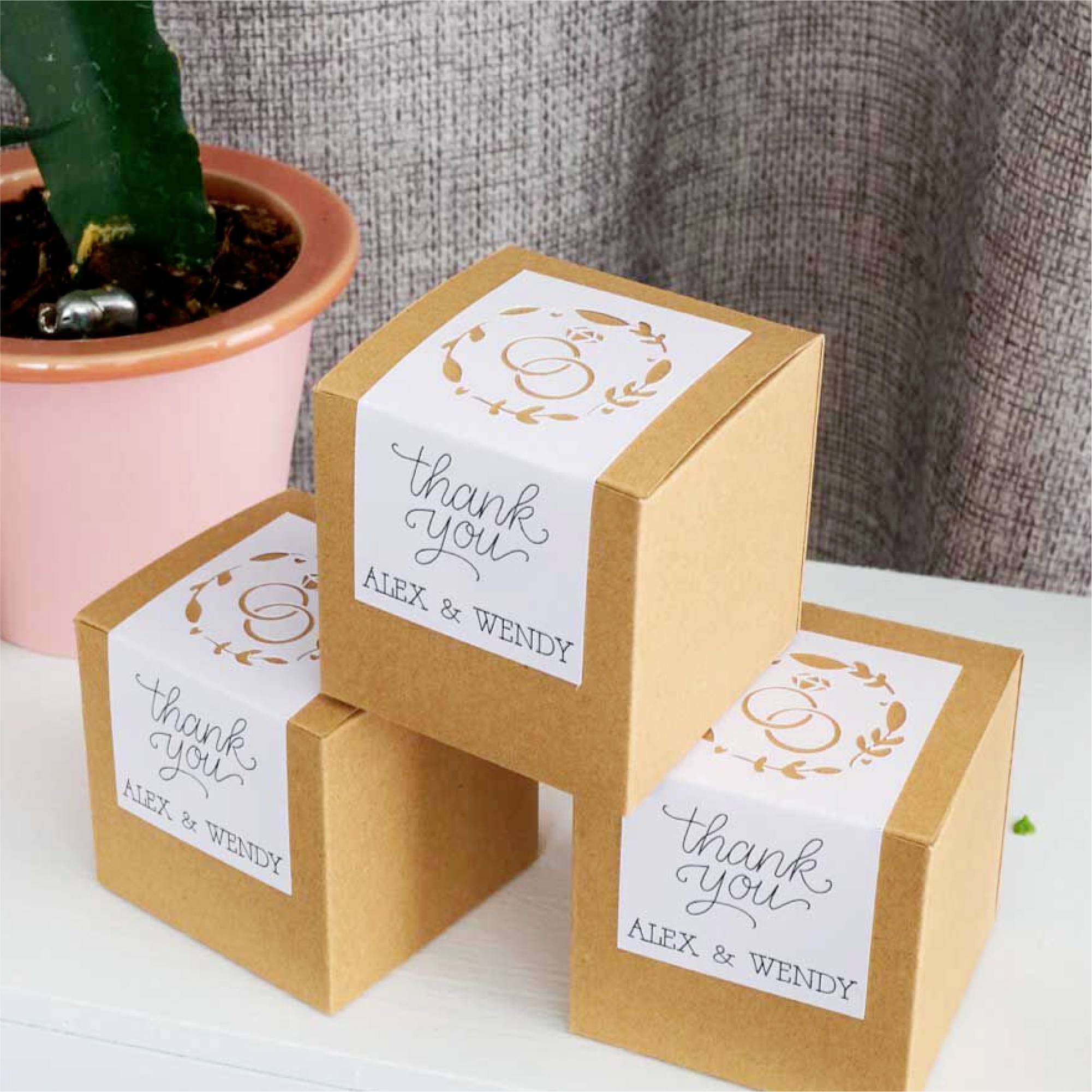 Drie bruine geschenkdozen met de tekst 'thank you Alex & Wendy' voor een pot met een plant, gerangschikt op een tafel.
