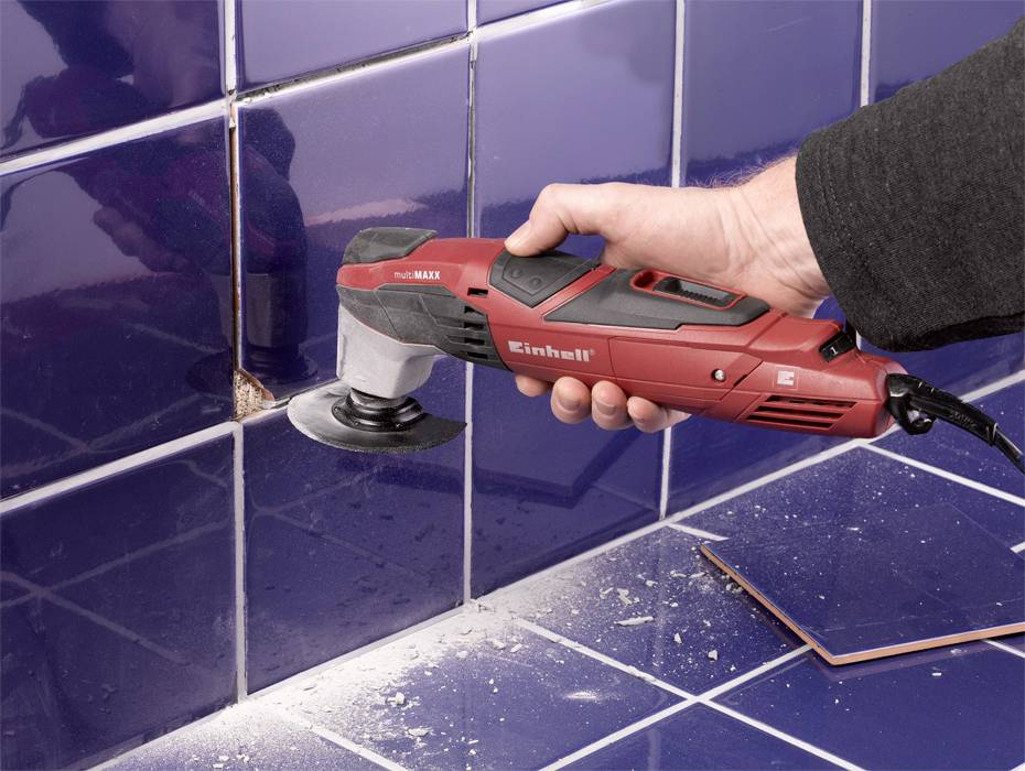 Een persoon verwijdert een beschadigde blauwe tegel met een rode elektrische multitool. Stof en brokstukken zijn zichtbaar.