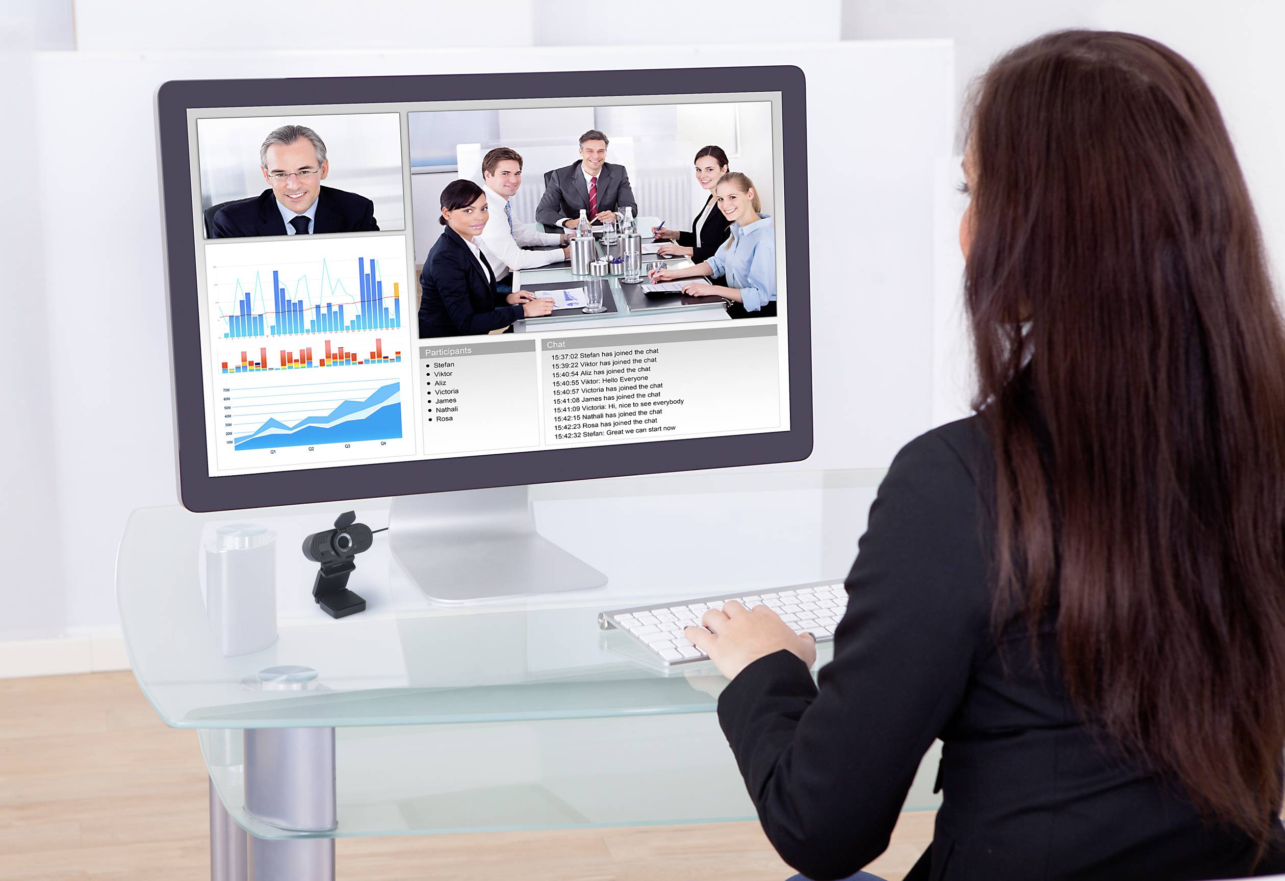 Een vrouw neemt deel aan een videoconferentie. Op het scherm zijn meerdere deelnemers, een diagram met staaf- en lijngrafieken, en tekst zichtbaar.