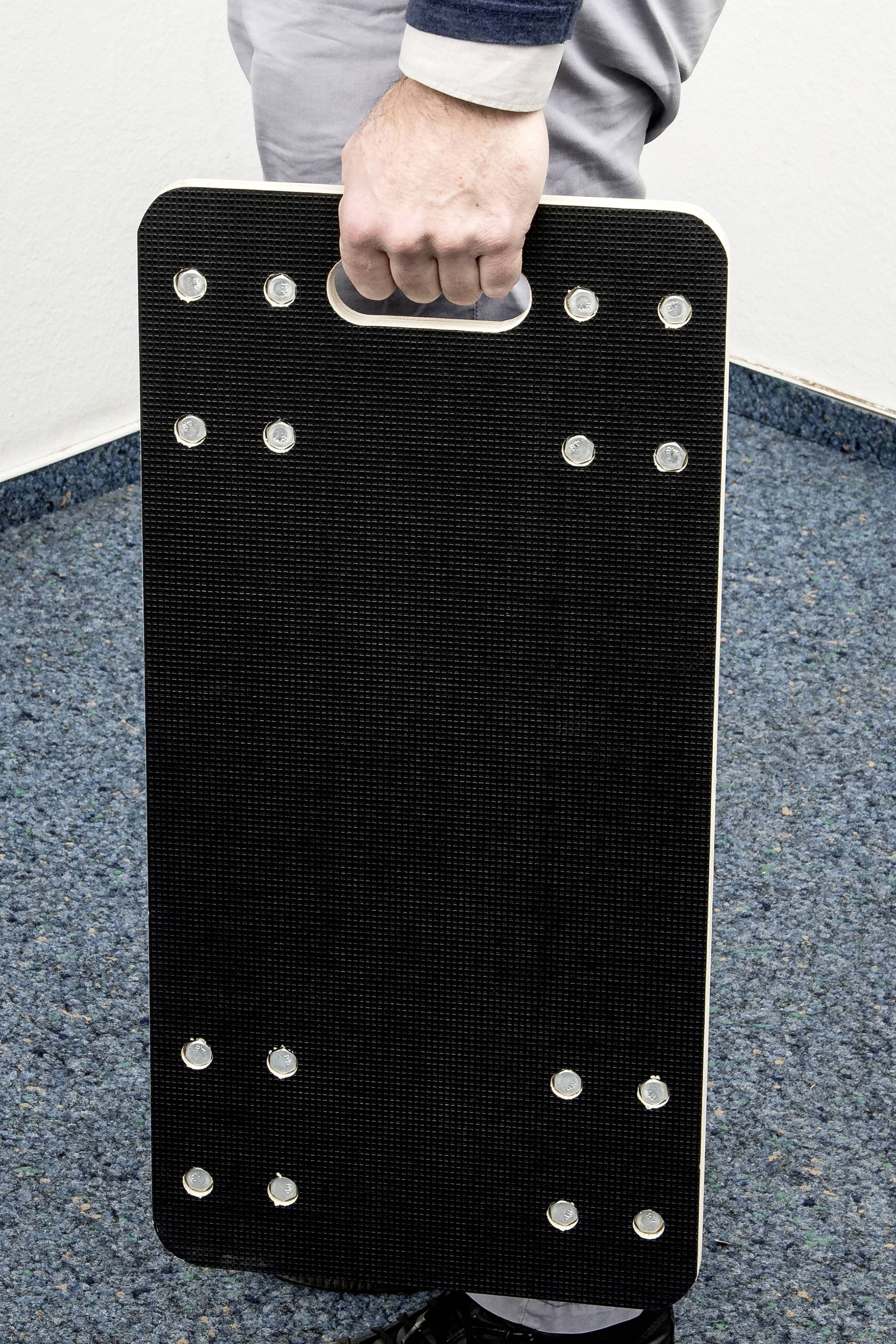 Een persoon houdt een rechthoekige, zwarte plaat met een handvat en meerdere bevestigingspunten, vermoedelijk een sporttoestel.