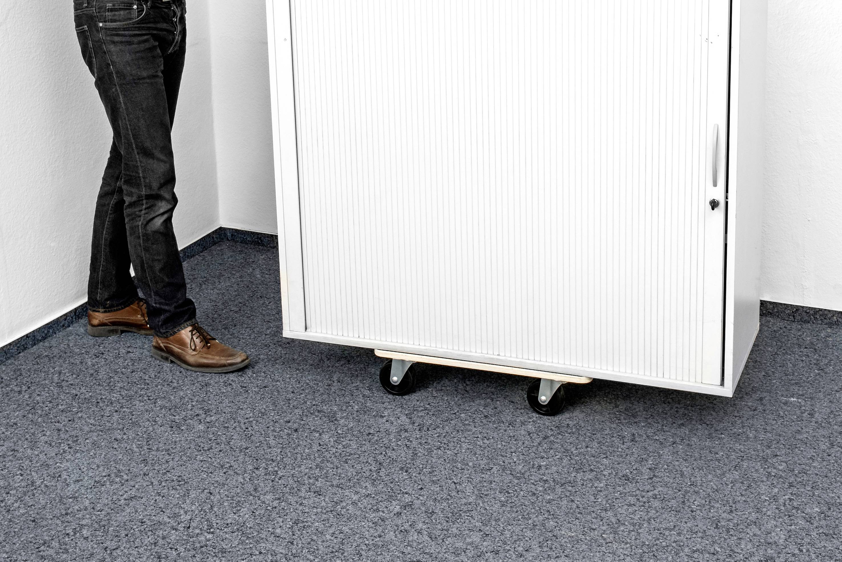Een persoon staat naast een witte afgesloten kast op wielen. De vloer is bedekt met grijze vloerbedekking.