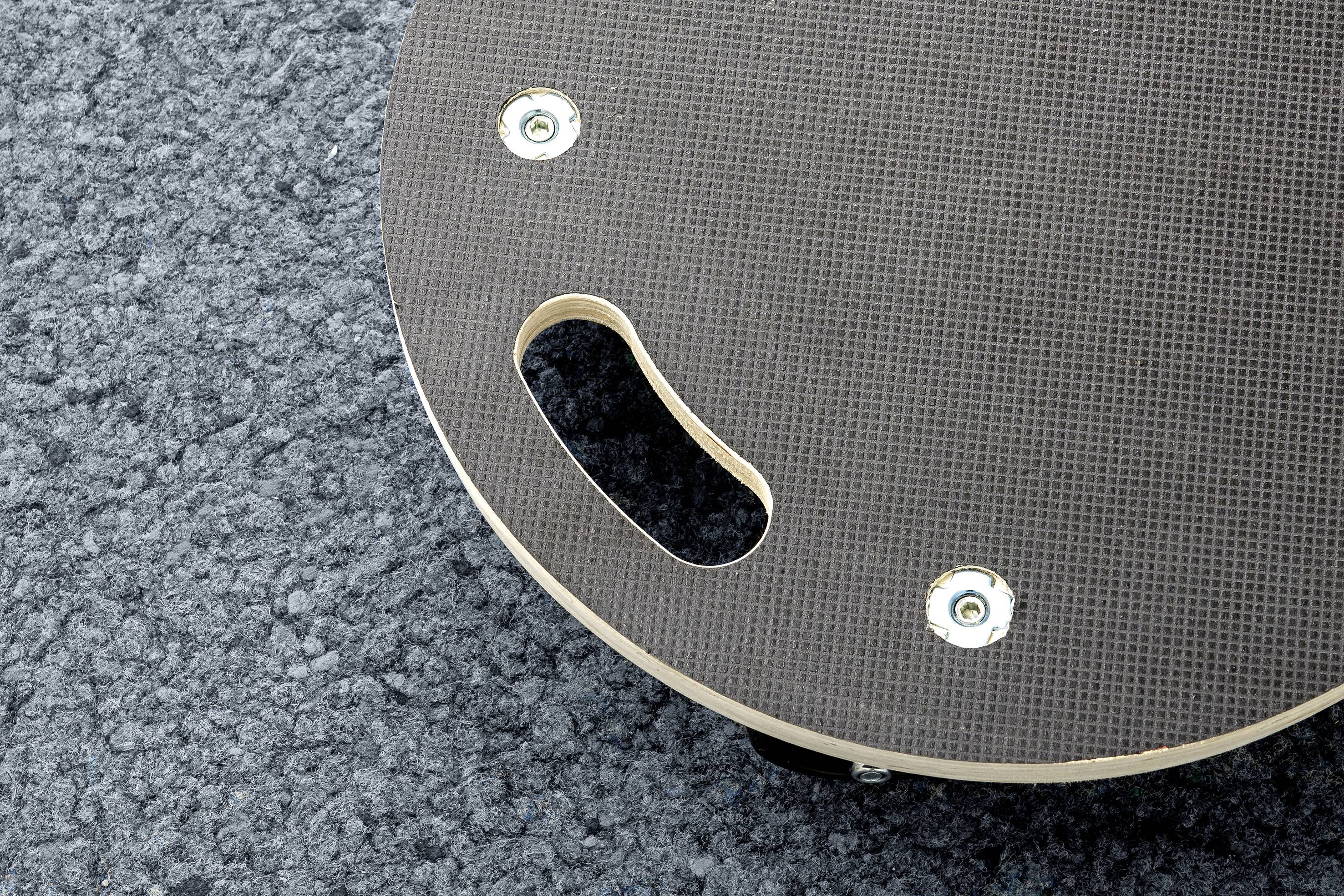 Een deel van een rond, grijs skateboard dat op een blauw-grijze vloerbedekking ligt. Twee schroeven en een handgreepgat zijn zichtbaar.