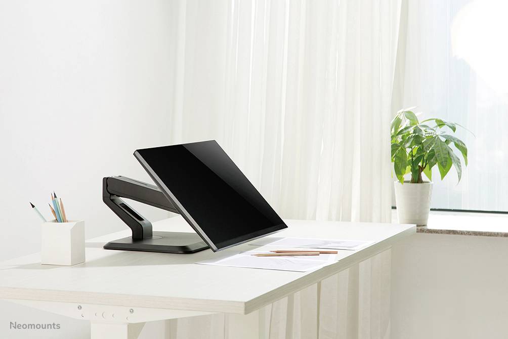 Een moderne bureau met een scherm op een verstelbare arm, naast een potje met pennen en een plant bij het raam.