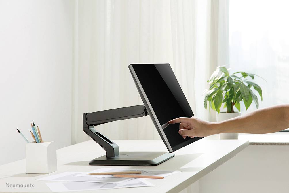 Een vinger raakt het scherm aan van een moderne, verstelbare monitor op een bureau naast schrijfbenodigdheden en een plant.