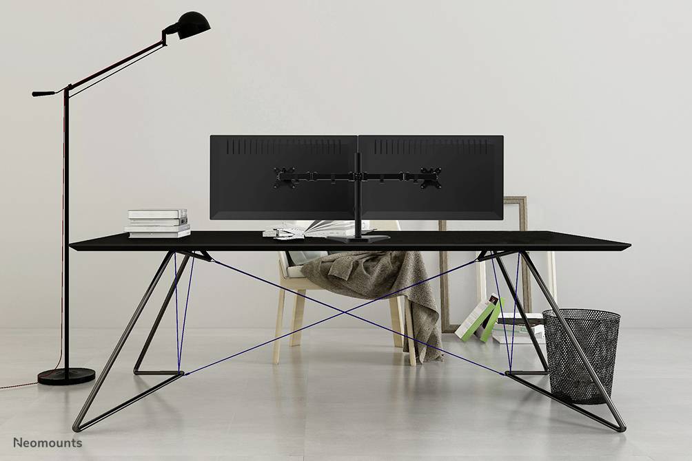Een moderne bureau met slanke metalen poten, daarop twee grote monitoren, ernaast een eenvoudige zwarte lampenkap. Op de achtergrond een stoel met een kussen.
