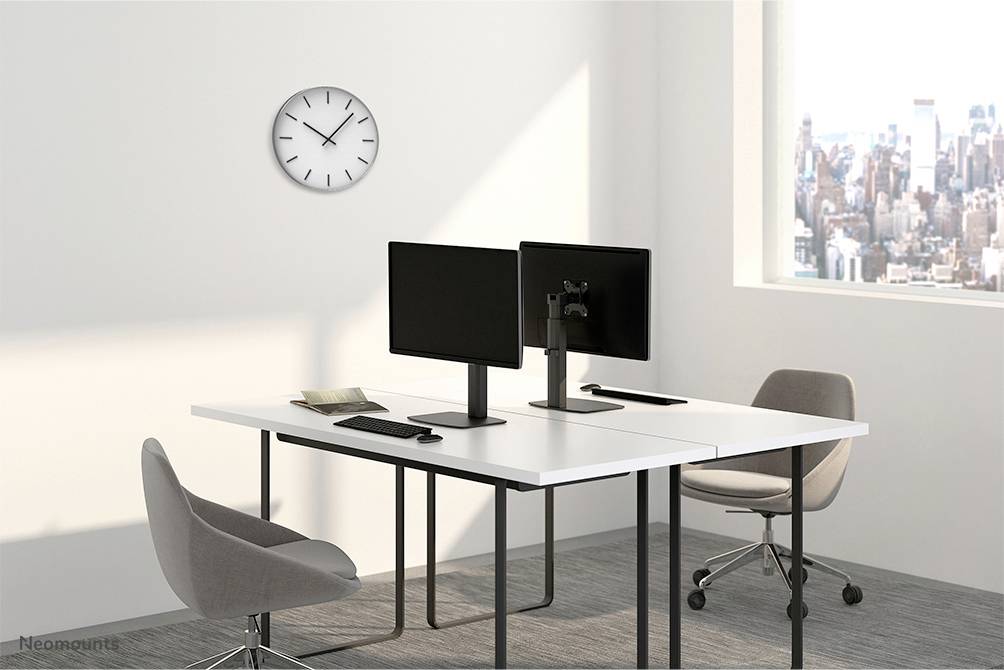 Een modern kantoor met twee computermonitoren op een witte tafel, twee grijze stoelen, een klok aan de muur en uitzicht op een stad door het raam.