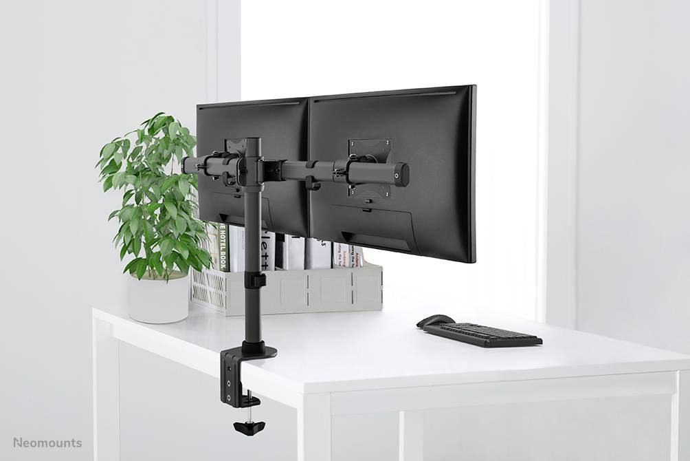 Een bureau met twee naast elkaar gemonteerde monitors, een toetsenbordset eronder, een plant links, in een lichte ruimte.