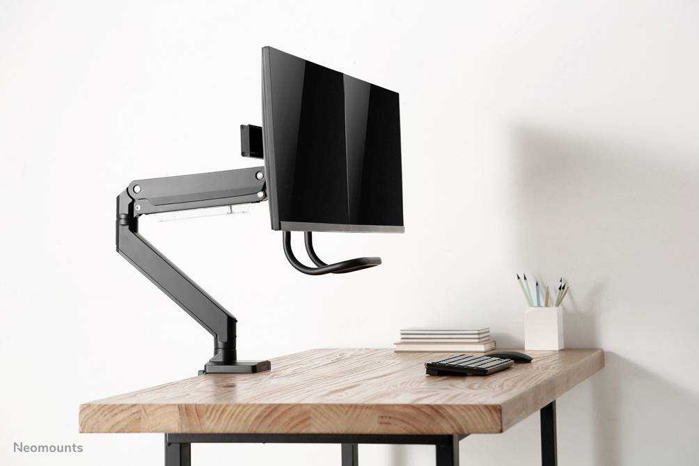 Een bureau met een gemonteerde monitorarm waaraan een computerscherm hangt; ernaast liggen pennen en notitieblokken.