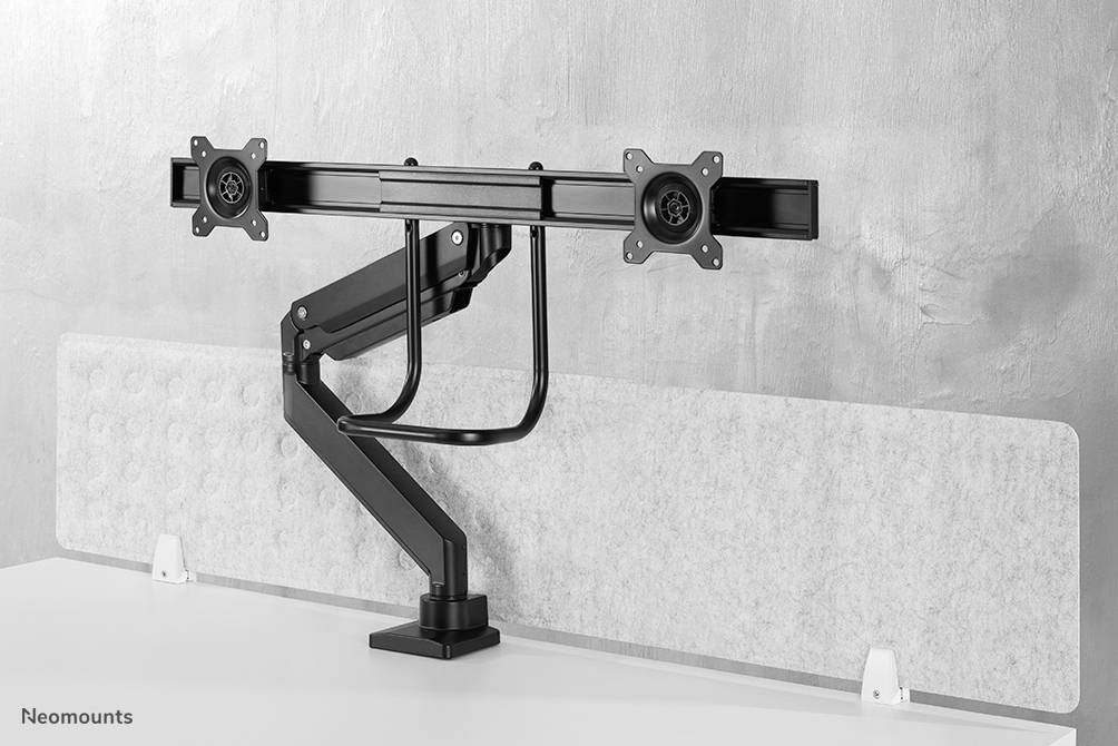 Dubbele monitorsteun met twee beweegbare armen, gemonteerd op een witte tafel tegen een grijze muur.