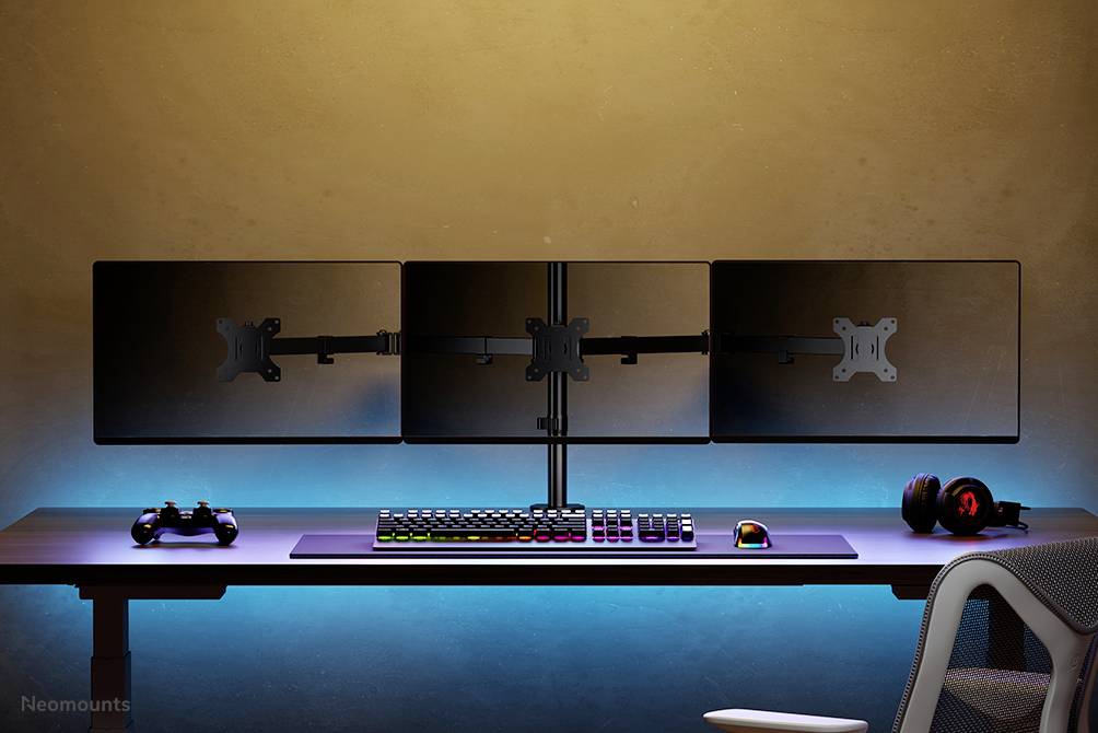 Drie monitors op een bureau met gaming-accessoires, waaronder toetsenbord, muis, headset en controller; blauwe LED-achtergrond.