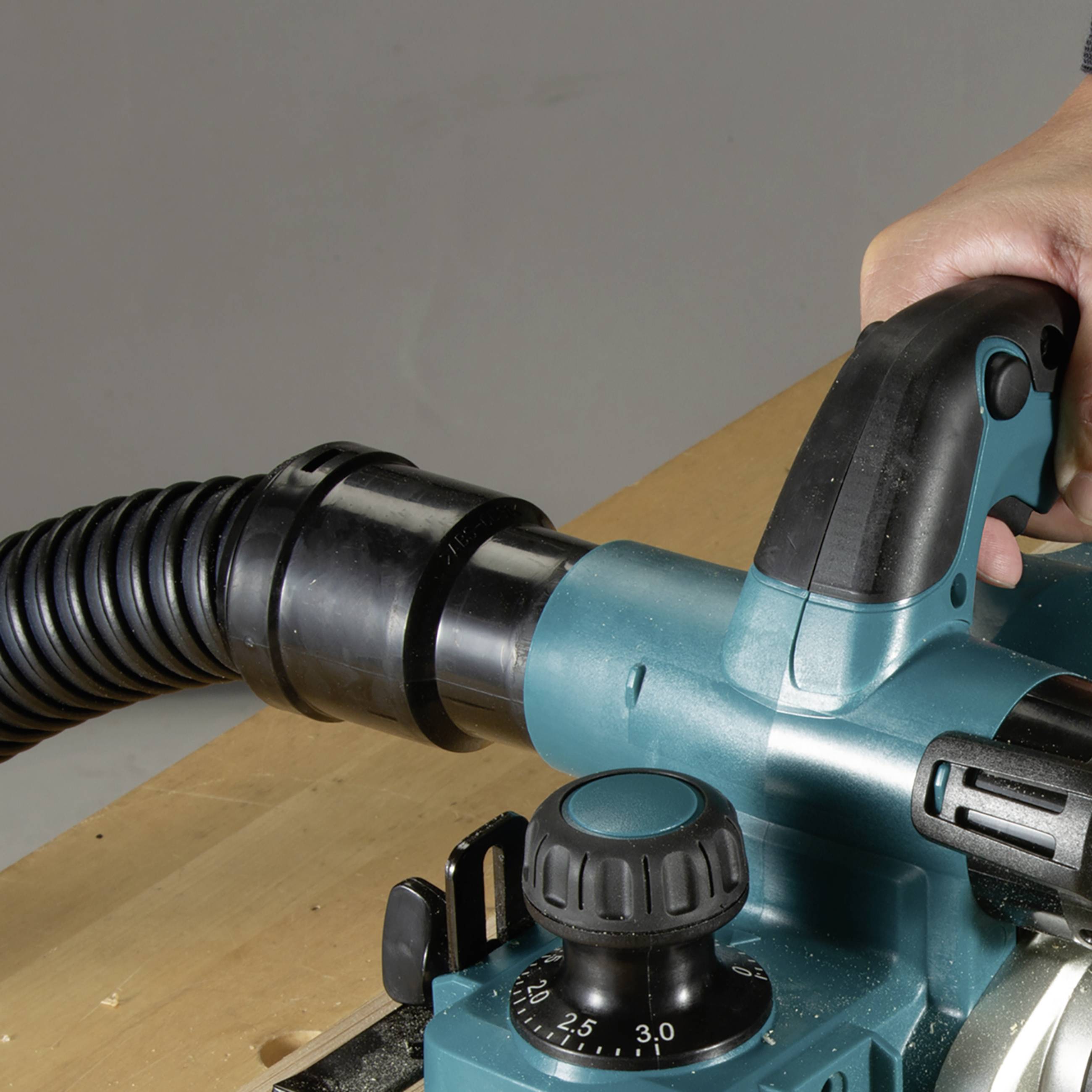 Een hand bedient een elektrische bovenfrees op een houten tafel. Een stofzuigerslang is op de machine aangesloten.