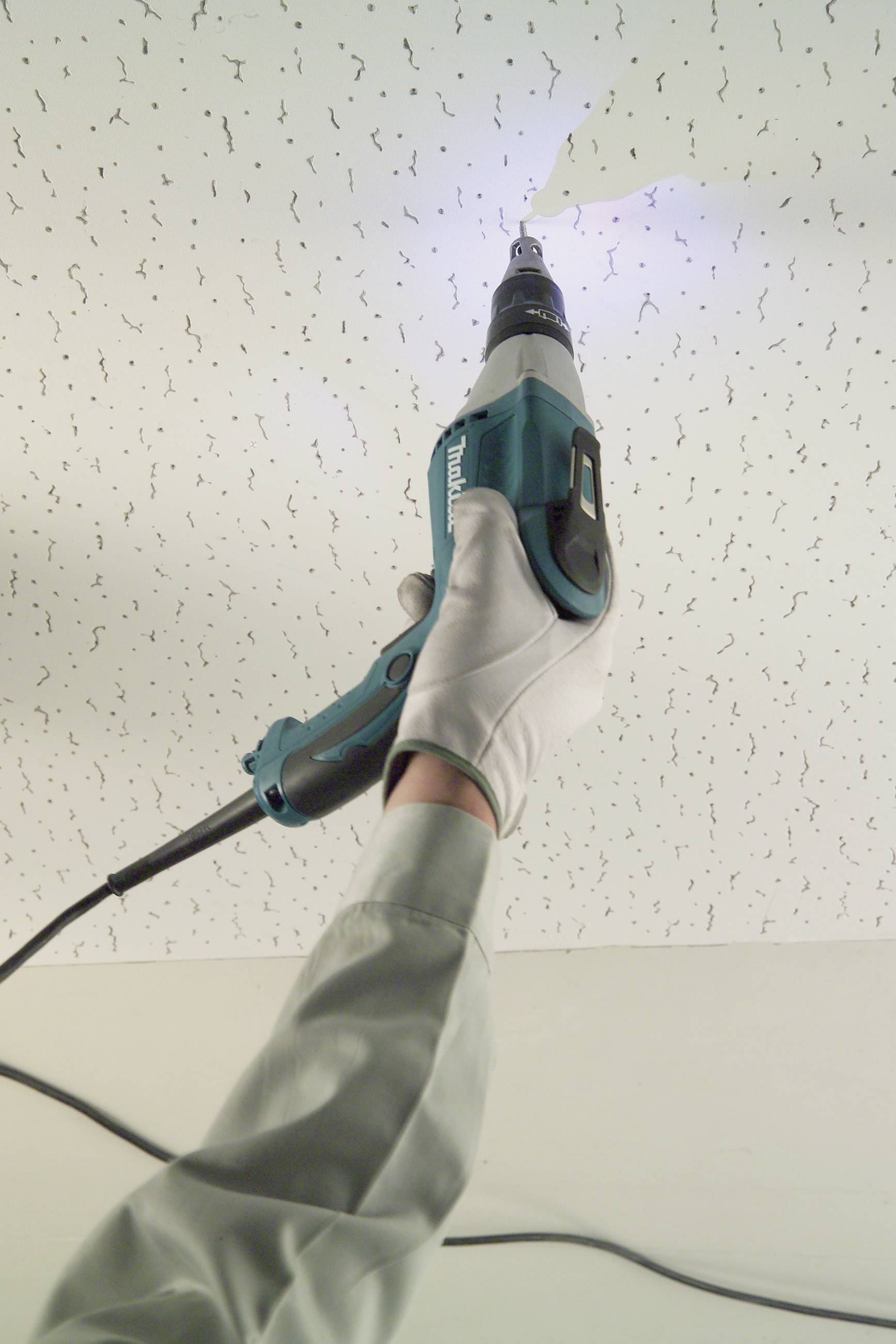 Een persoon boort met een blauwe elektrische schroevendraaier een gat in een wit plafond. De hand draagt een witte handschoen.
