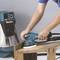 Een persoon slijpt een houten tafel met een blauwe elektrische slijpmachine. Op de achtergrond staat een blauwe stofzuiger.