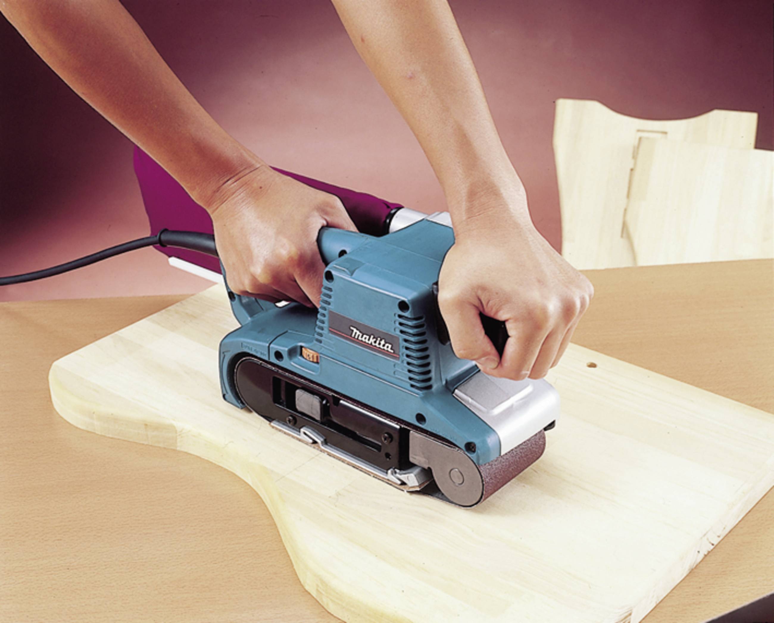 Een persoon schuurt een houten plank met een elektrische bandschuurmachine. Het apparaat is blauw en staat op een bruine tafel.