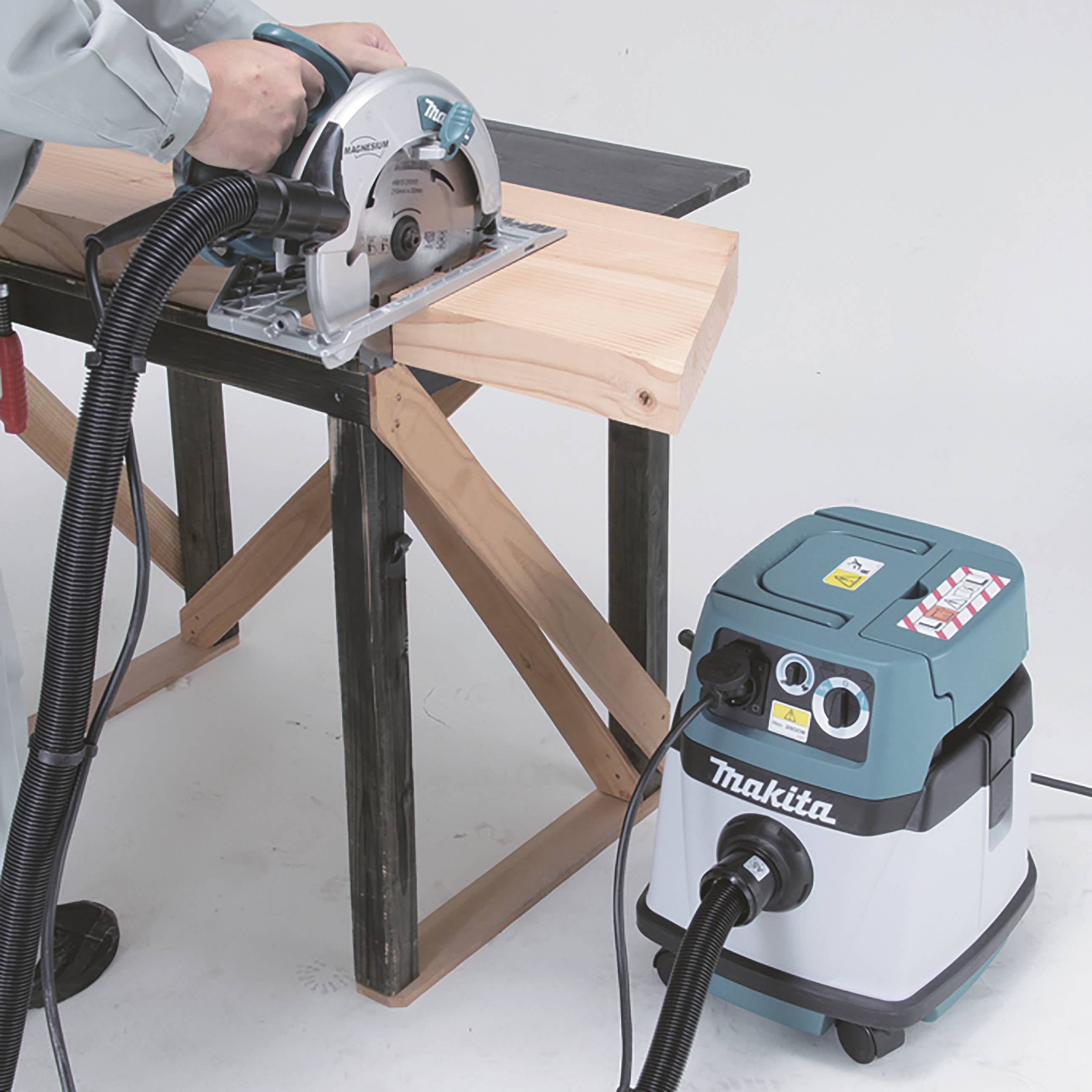 Een persoon zaagt een houten plank door met een cirkelzaag aan een tafel. Een aangesloten stofzuiger van Makita zuigt houtkrullen op.