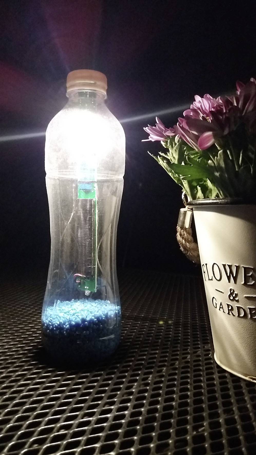 Een plastic fles met blauw granulaat en een markeerstift vanbinnen licht op aan een tafel naast een bloempot met roze bloemen.