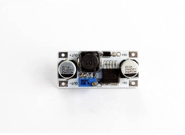 Een Spaans spanningsregelmodule met twee condensatoren, een spoelkern en aansluitingen, gemarkeerd met 'IN+' en 'OUT-'.