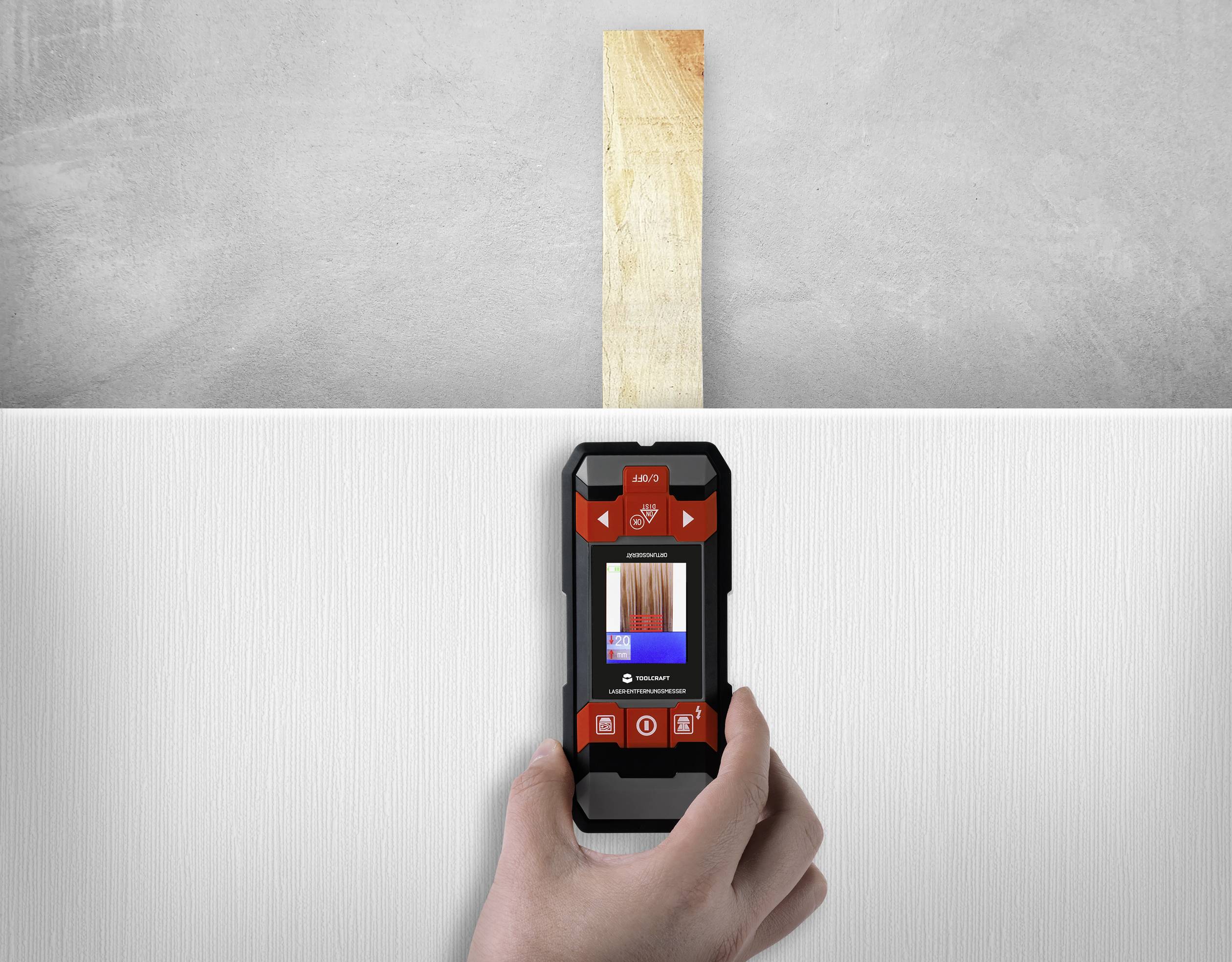 Een hand houdt een vochtmeter voor hout boven een houten plank op een witte achtergrond. Het apparaat toont meetresultaten.