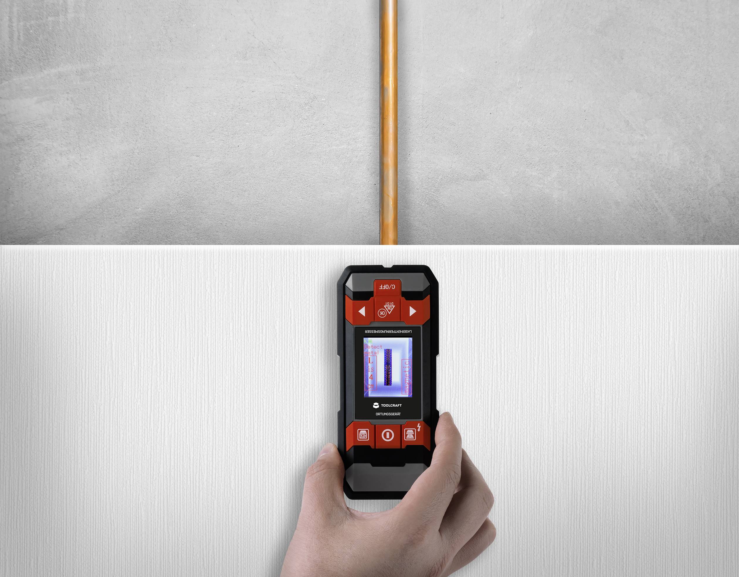 Een hand houdt een digitaal opsporingsapparaat vast dat een verticaal lopende leiding in de muur detecteert.