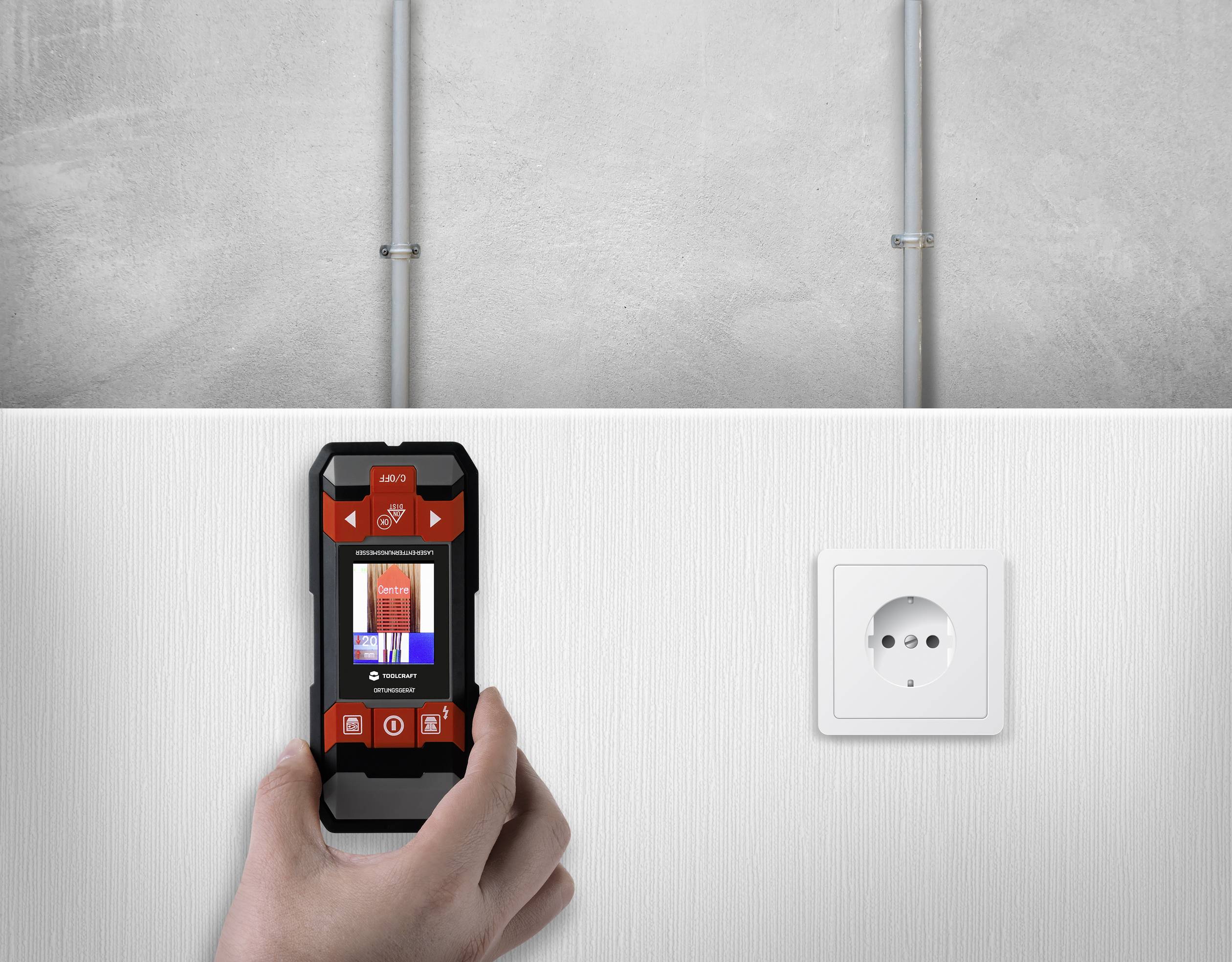 Een hand houdt een muurscanner voor een witte muur. Op het scherm van de scanner zijn rode markeringen zichtbaar. Rechts bevindt zich een stopcontact.