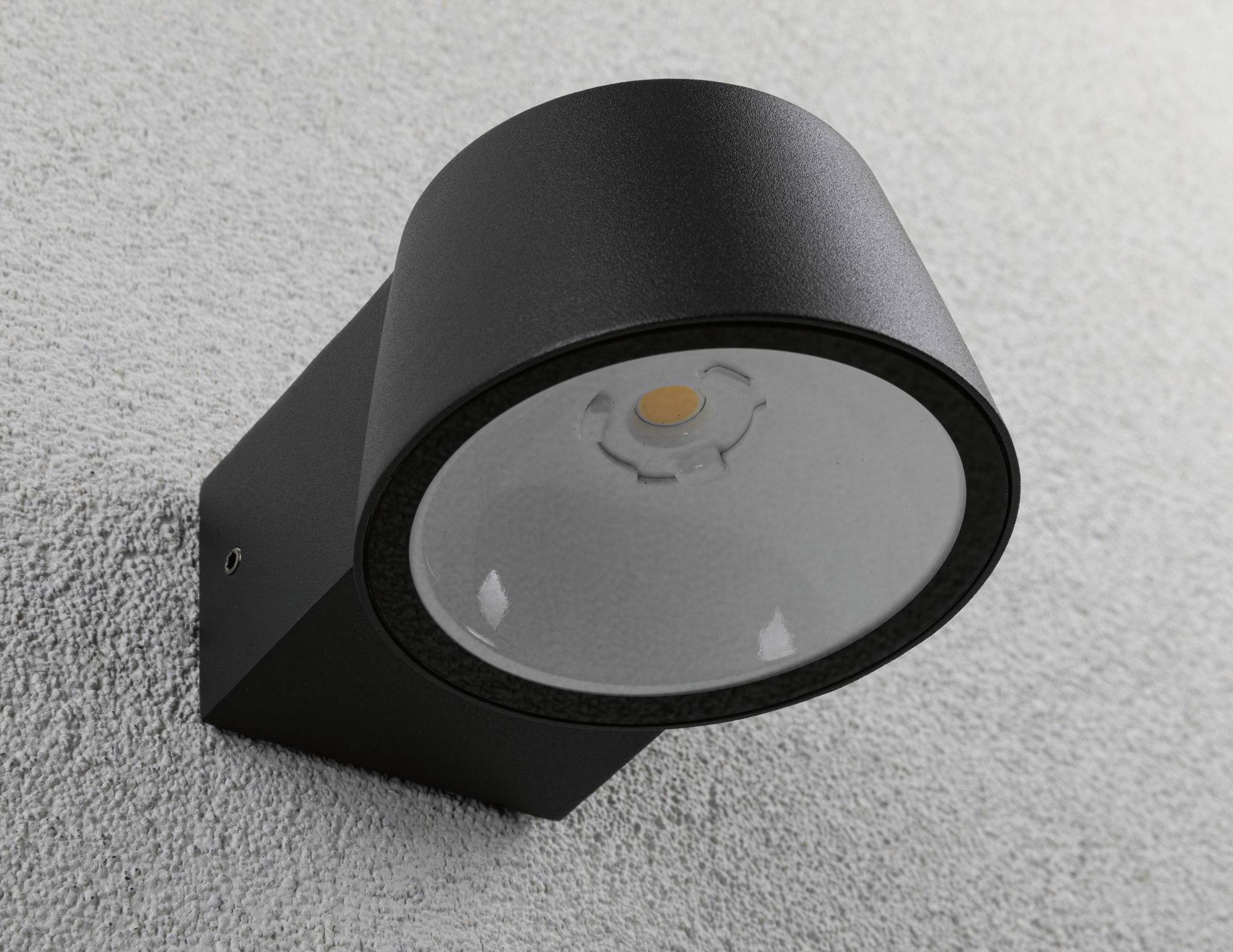 Zwarte, cilindrische buitenlamp gemonteerd aan een gestructureerde grijze muur, met een LED-verlichting aan de binnenkant.