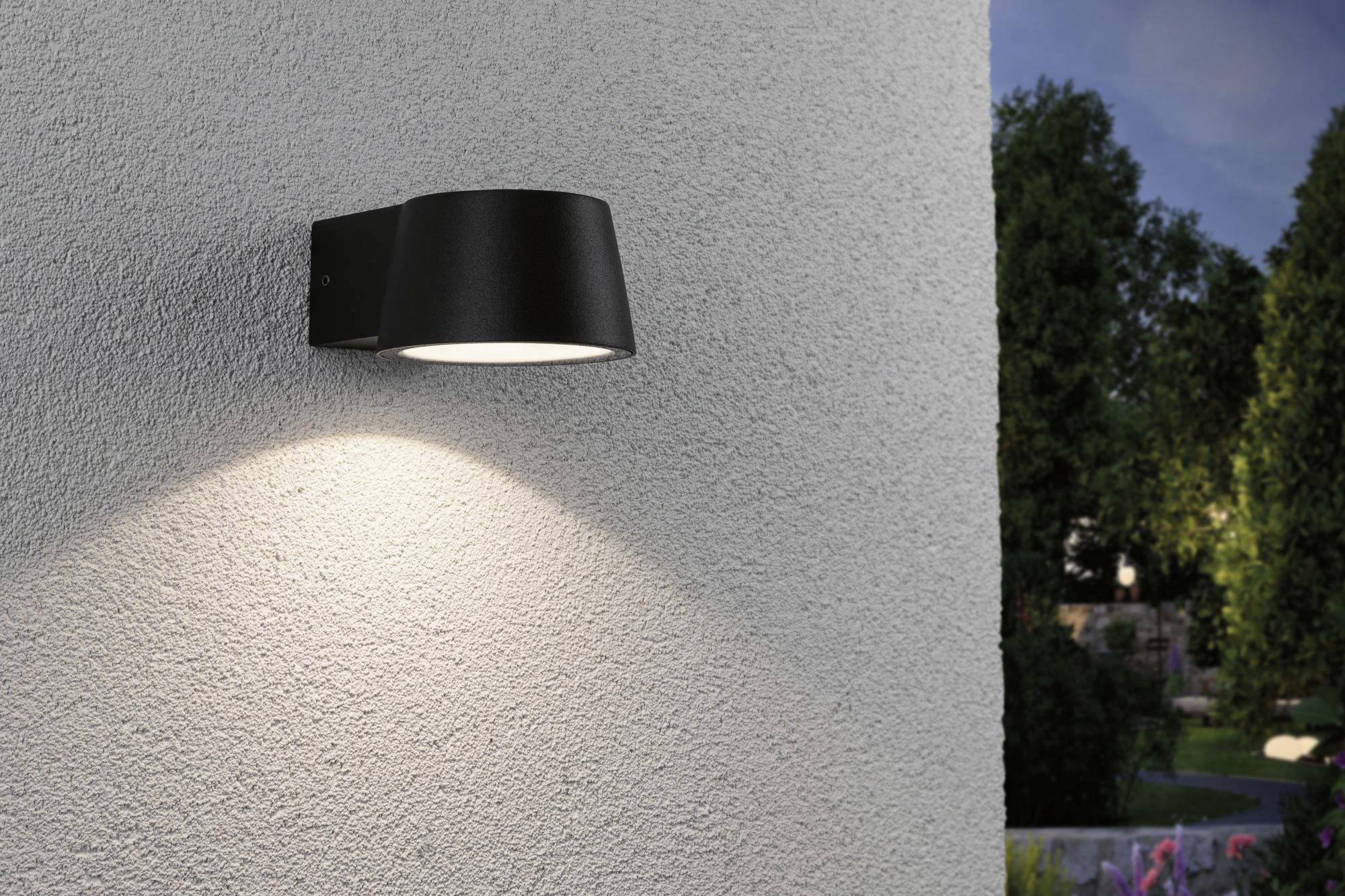 Zwarte buitenwandlamp aan grijze muur schijnt naar beneden en verlicht een bepaald gebied. Op de achtergrond is een tuin zichtbaar.