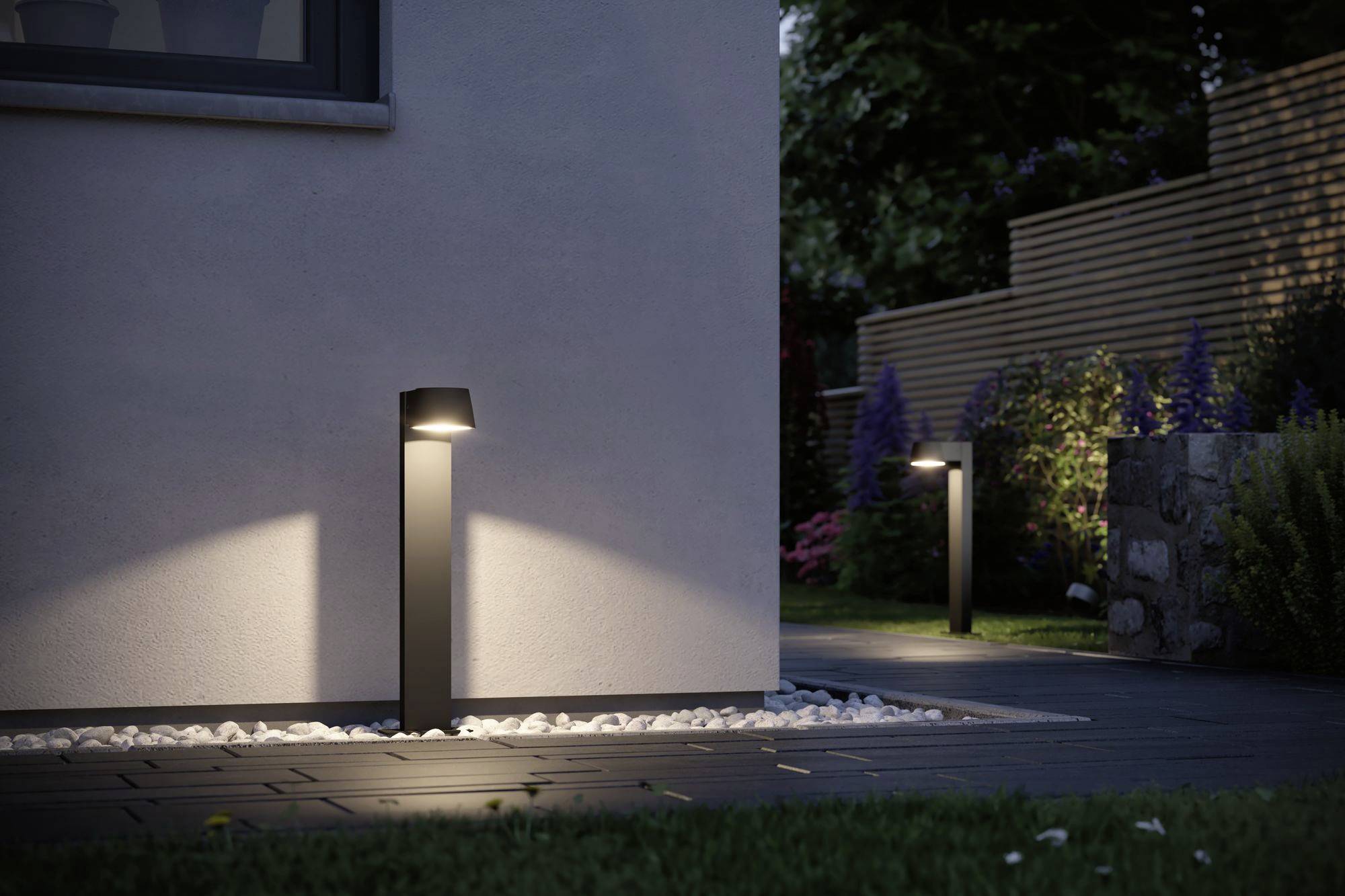 Tuinverlichting bij schemering: Twee moderne lampen verlichten een bestrate weg naast een huis, omgeven door planten.