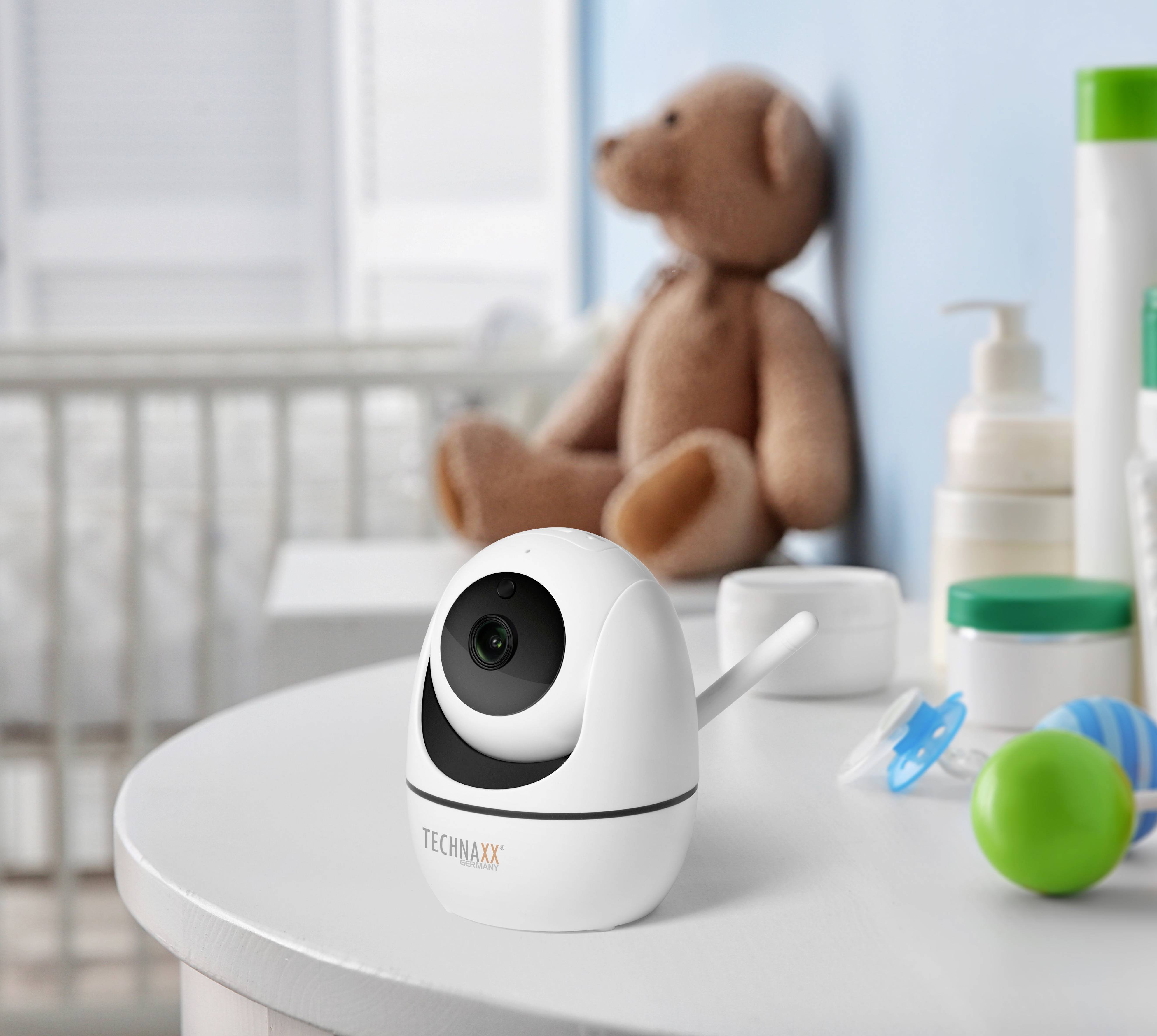 Een babycamera staat op een tafel in de kinderkamer, naast speelgoed en verzorgingsproducten. Op de achtergrond een teddybeer en een kinderbed.