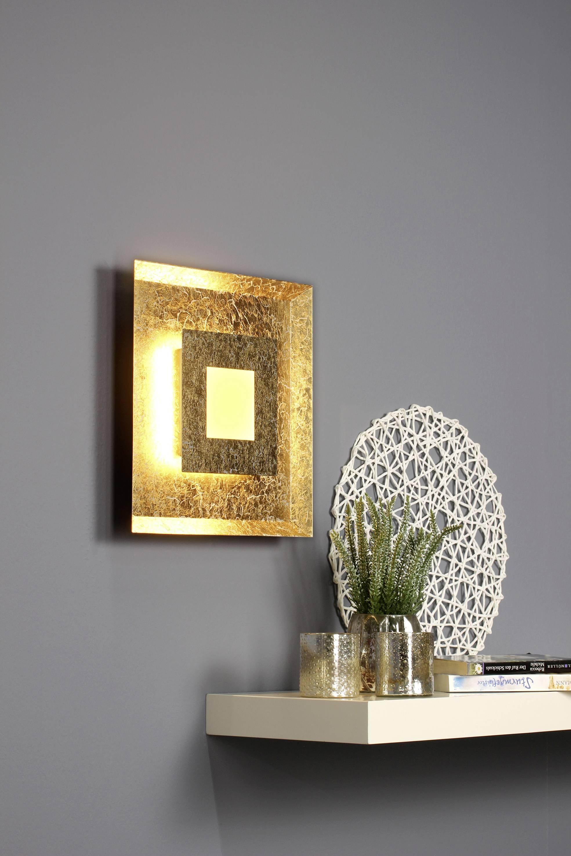 Een stralende, vierkante wandspiegel met gouden details. Eronder een plank met een plant, een kandelaar en boeken op een grijze muur.