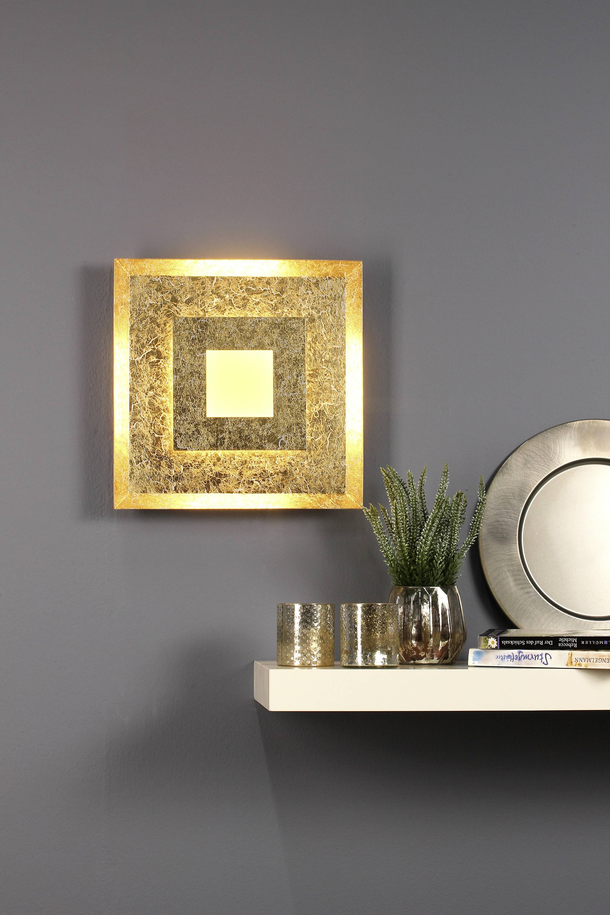 Wanddecoratie met goud-zwart vierkant design schittert. Eronder een rek met een plant, kandelaar en een stapel boeken.