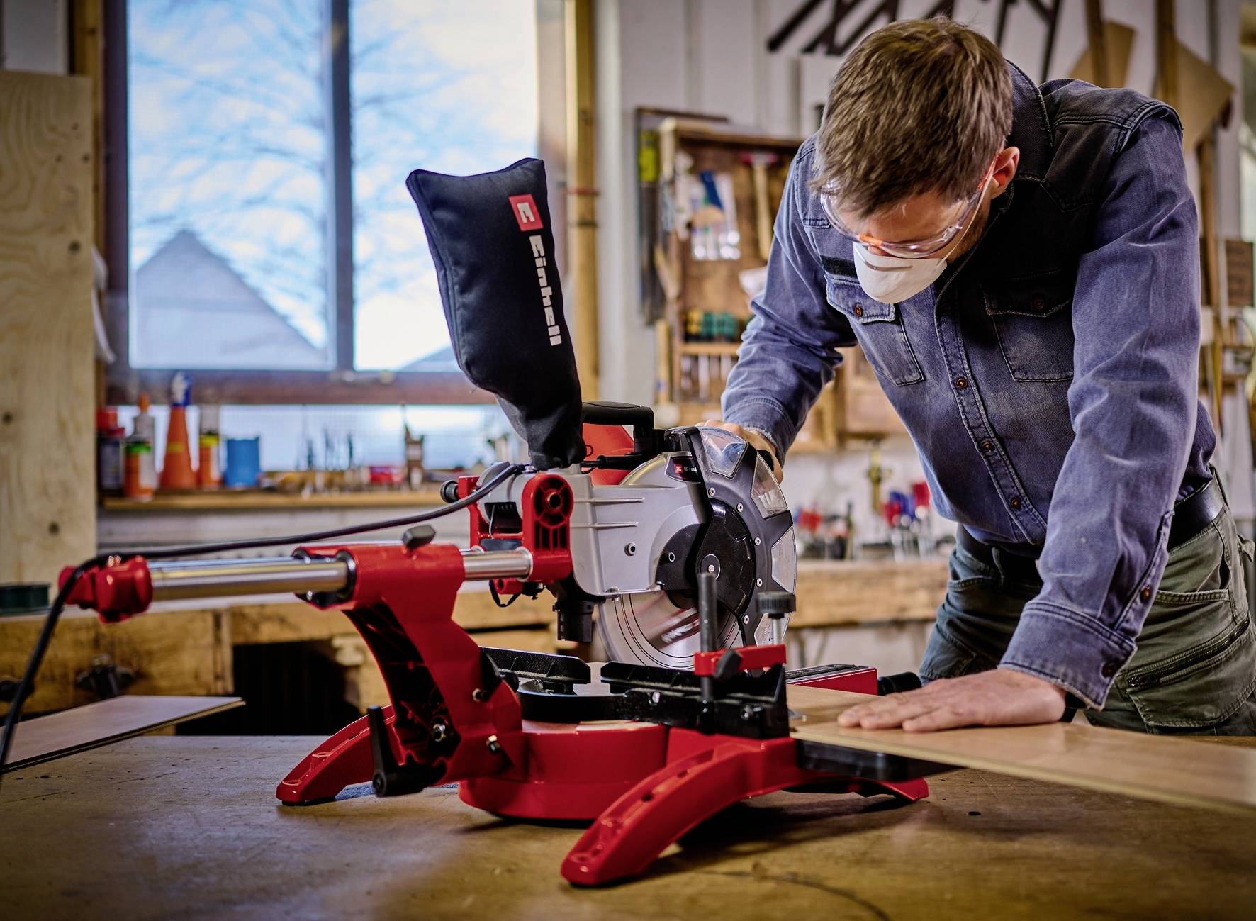 Een man met veiligheidsbril en masker zaagt een houten plank met een rode verstekzaag in een werkplaats.