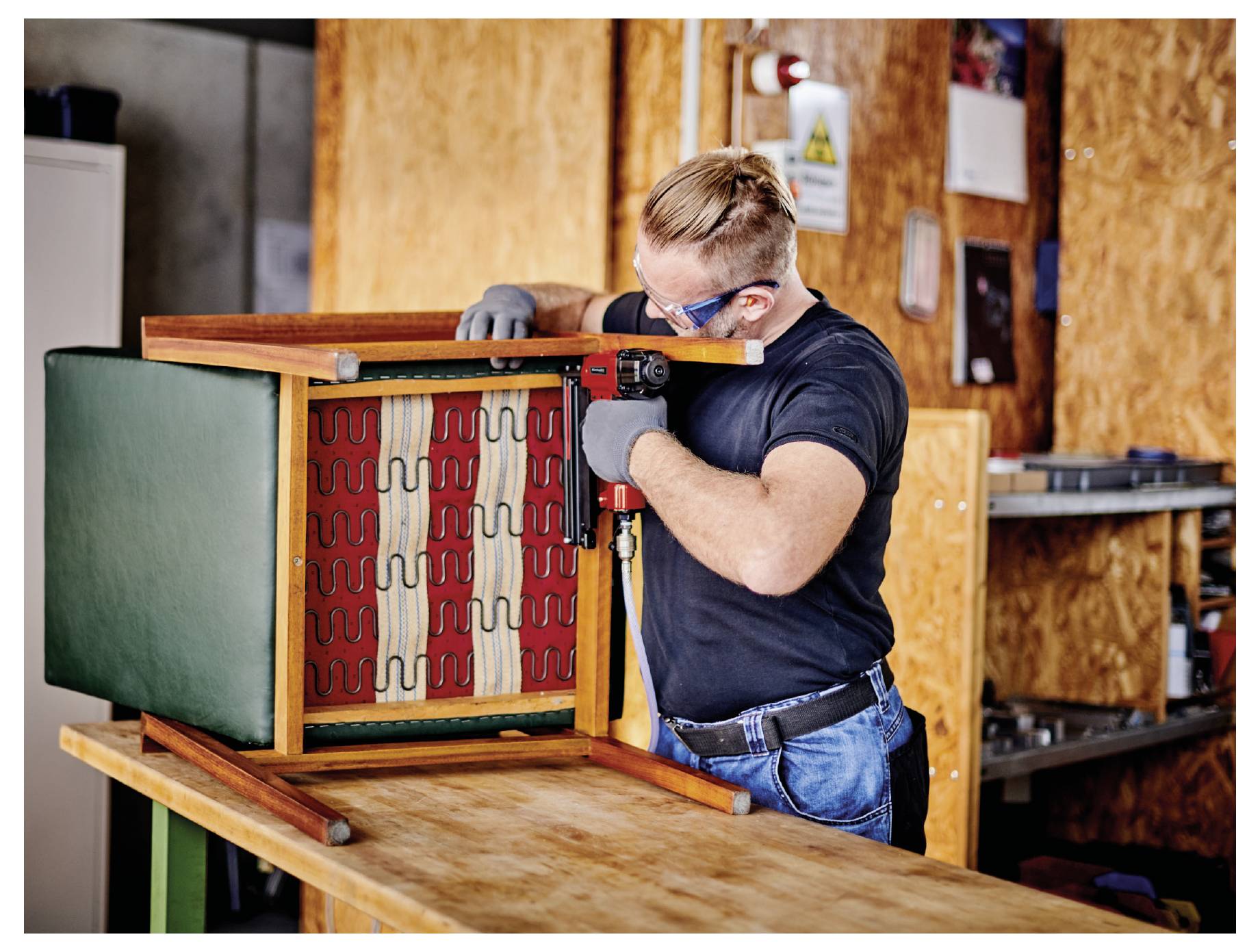 Man die een gestoffeerde stoel met een boormachine in een werkplaats repareert, gebogen over het omgekeerde frame op een werkbank.