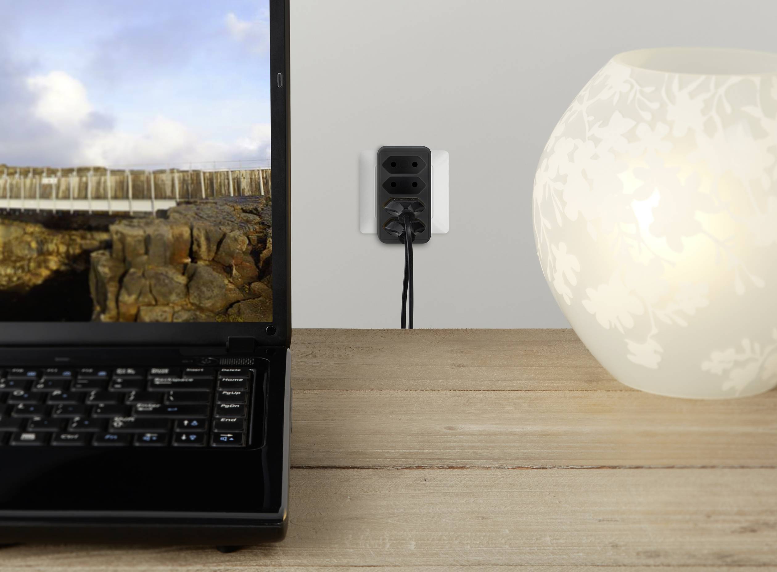 Een laptop toont een landschap, ernaast een verlichte lamp op een houten tafel. Op de achtergrond een stopcontact met een ingestoken kabel.