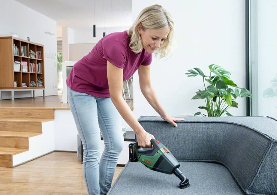 Een vrouw gebruikt een handstofzuiger om een grijze bank in een moderne woonkamer schoon te maken. Links zijn planken te zien.