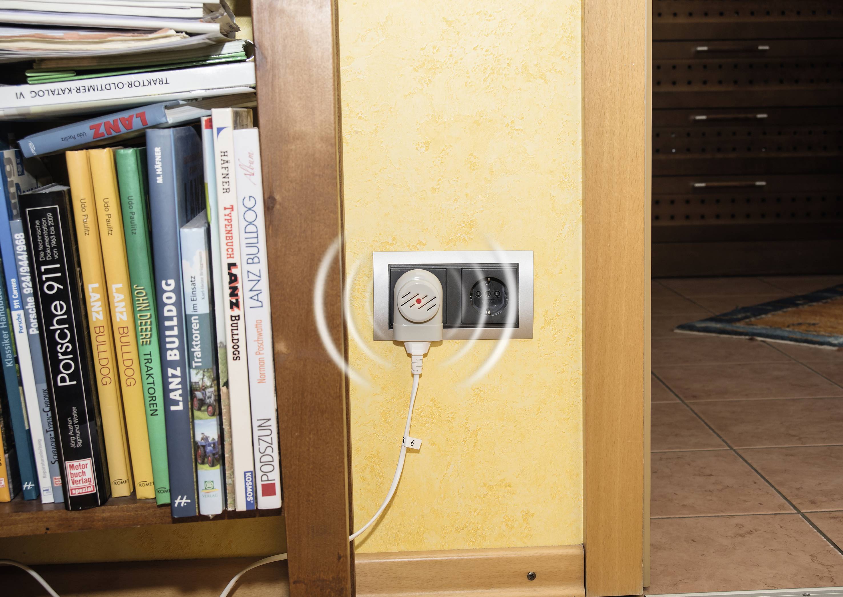 Boekenkast vol boeken naast een wandcontactdoos waarop een elektrisch apparaat is aangesloten.