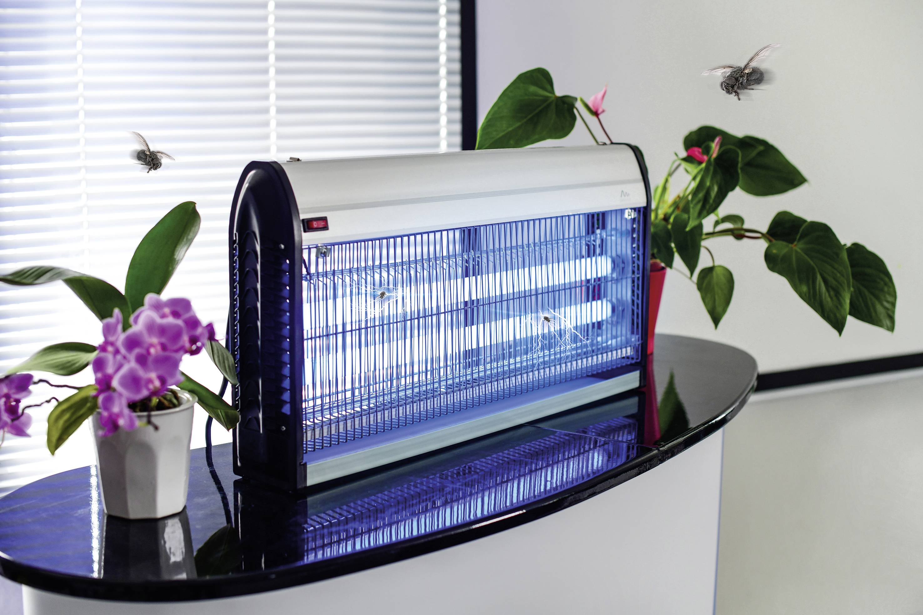 Elektrische insectenverdelger op een tafel naast planten, omringd door vliegende insecten, in een helder verlichte ruimte.