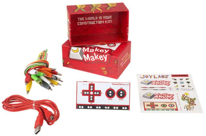 Makey Makey elektronische set met kabels en aansluitbladen. Verpakking op de achtergrond draagt de tekst 'De wereld is jouw constructieset!'